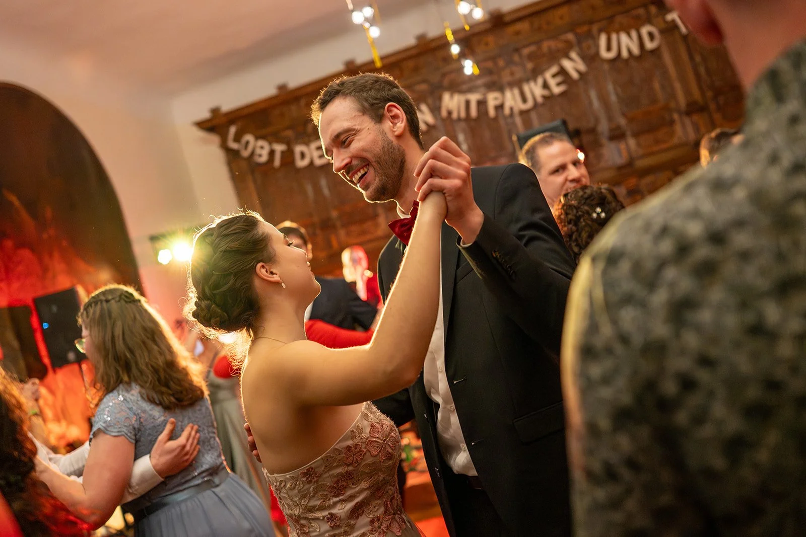 Ein Paar tanzt auf einer Hochzeitsfeier, das Mädchen trägt ein rotes Kleid und der Mann einen schwarzen Anzug. Im Hintergrund sind andere Gäste und eine Holzverzierung mit einem Spruch auf Deutsch.