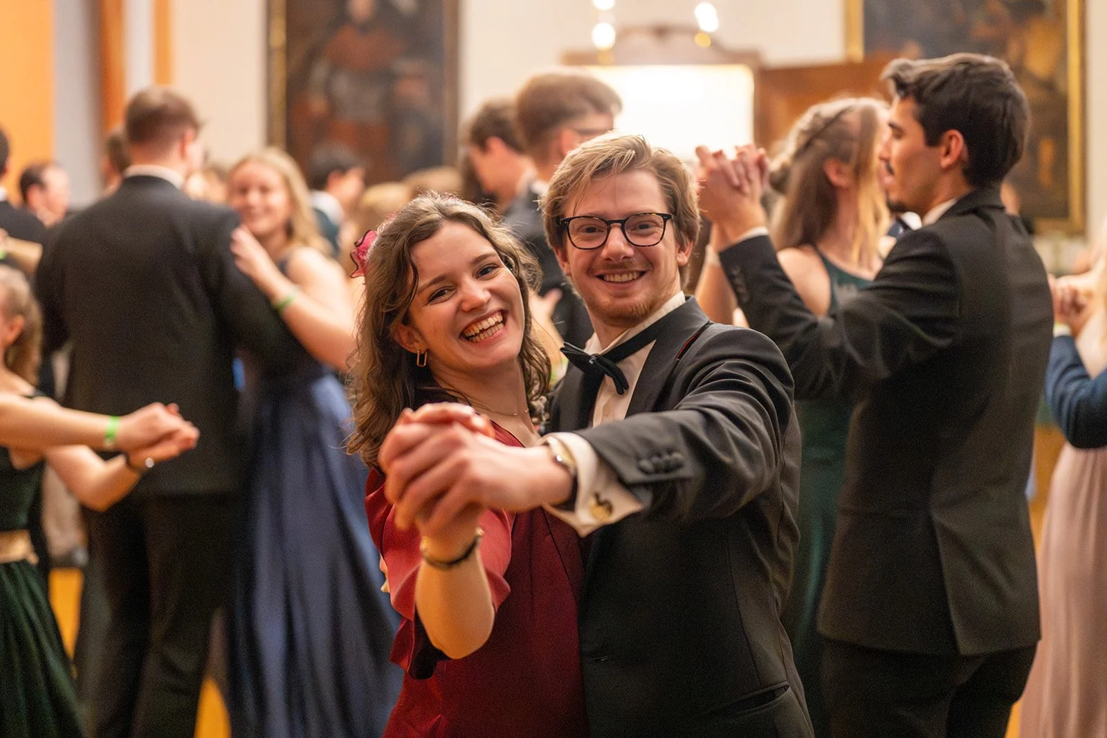 Tanzende Paare auf einer festlichen Veranstaltung, im Vordergrund ein lachendes Paar, das sich beim Tanzen hält, alle in eleganter Kleidung.