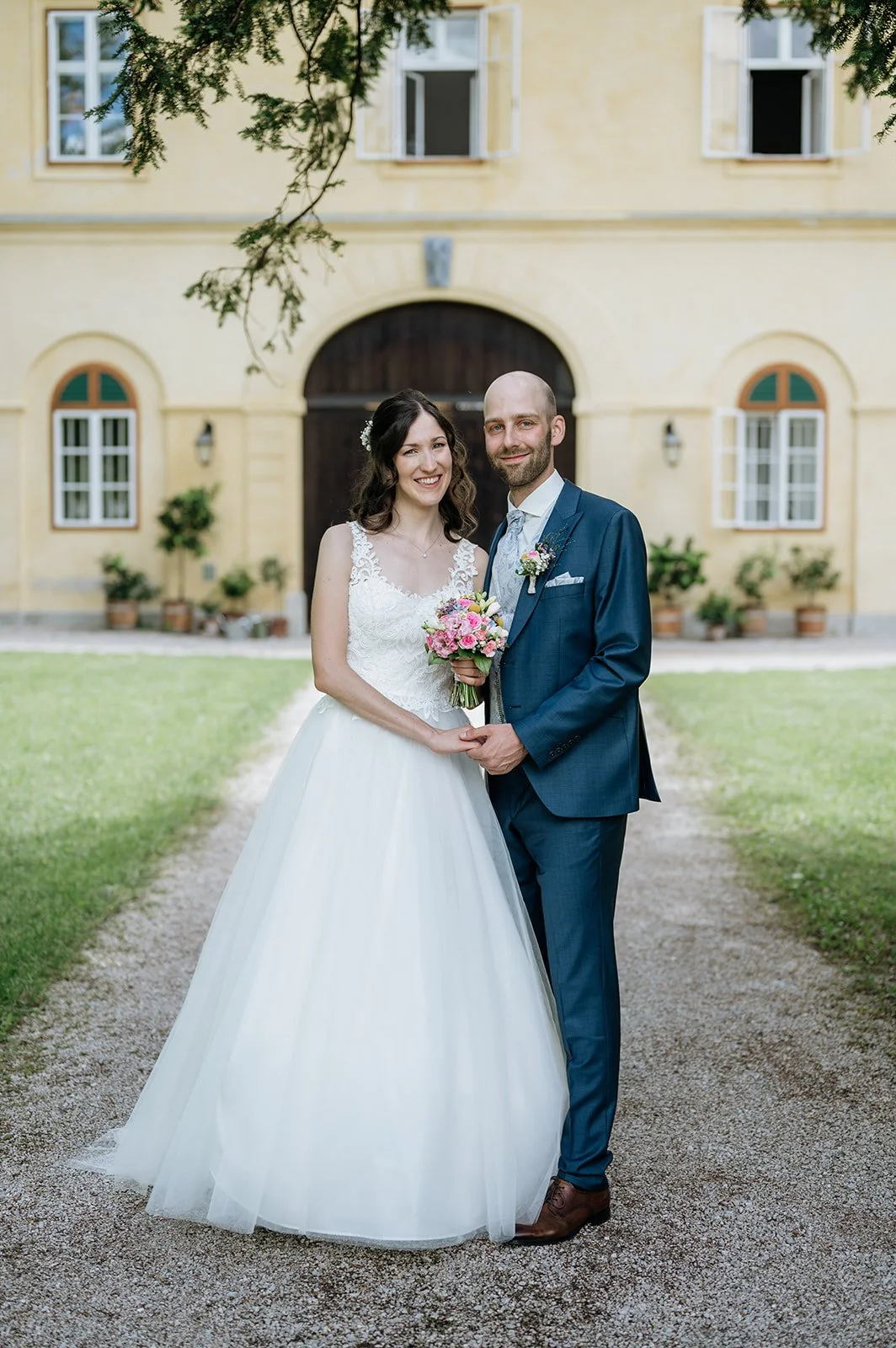 Ein Brautpaar vor einem gelben Gebäude, die Braut trägt ein weißes Hochzeitskleid mit Spitzenärmel und hält einen Blumenstrauß, der Bräutigam trägt einen blauen Anzug mit Krawatte und Fliege.
