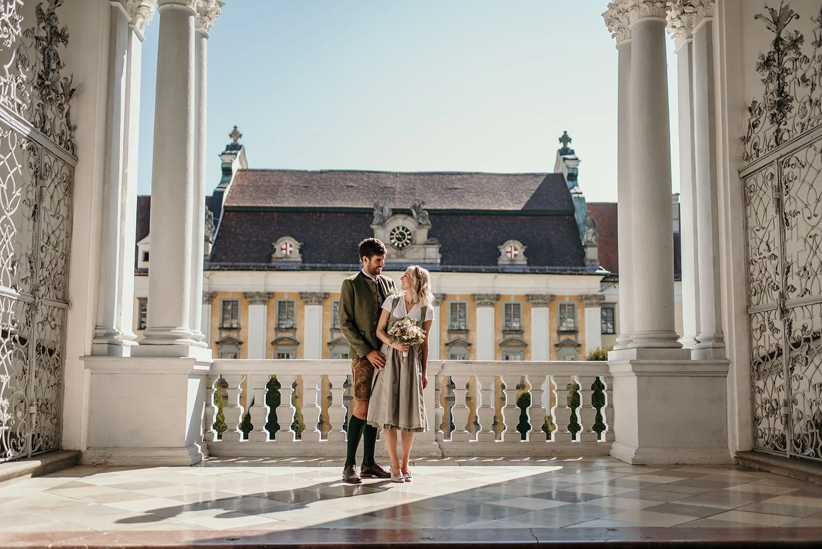 Ein Paar in traditionellen Trachten auf einer Porzellanveranda vor einem mittelalterlichen Gebäude, vermutlich bei einer Hochzeit im alpinen Raum.