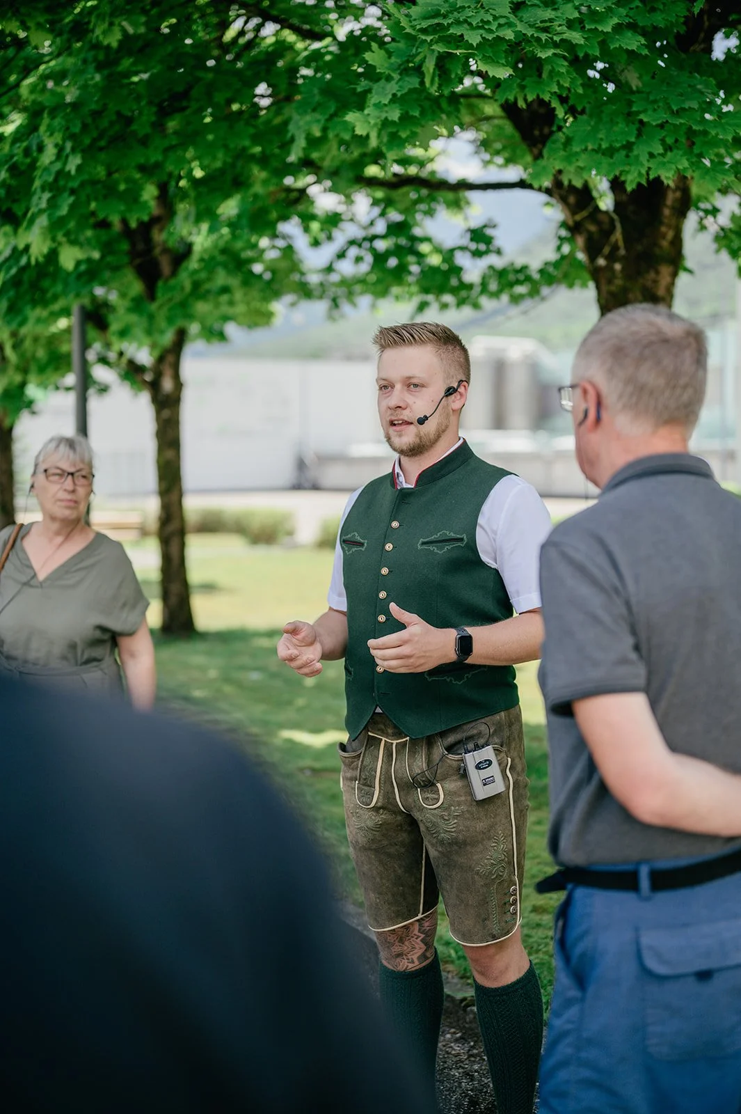 Ein junger Mann in traditioneller bayerischer Tracht spricht bei einer Veranstaltung im Freien, während zwei ältere Männer und eine Frau zuhören. Es gibt Bäume und eine Industrieanlage im Hintergrund.