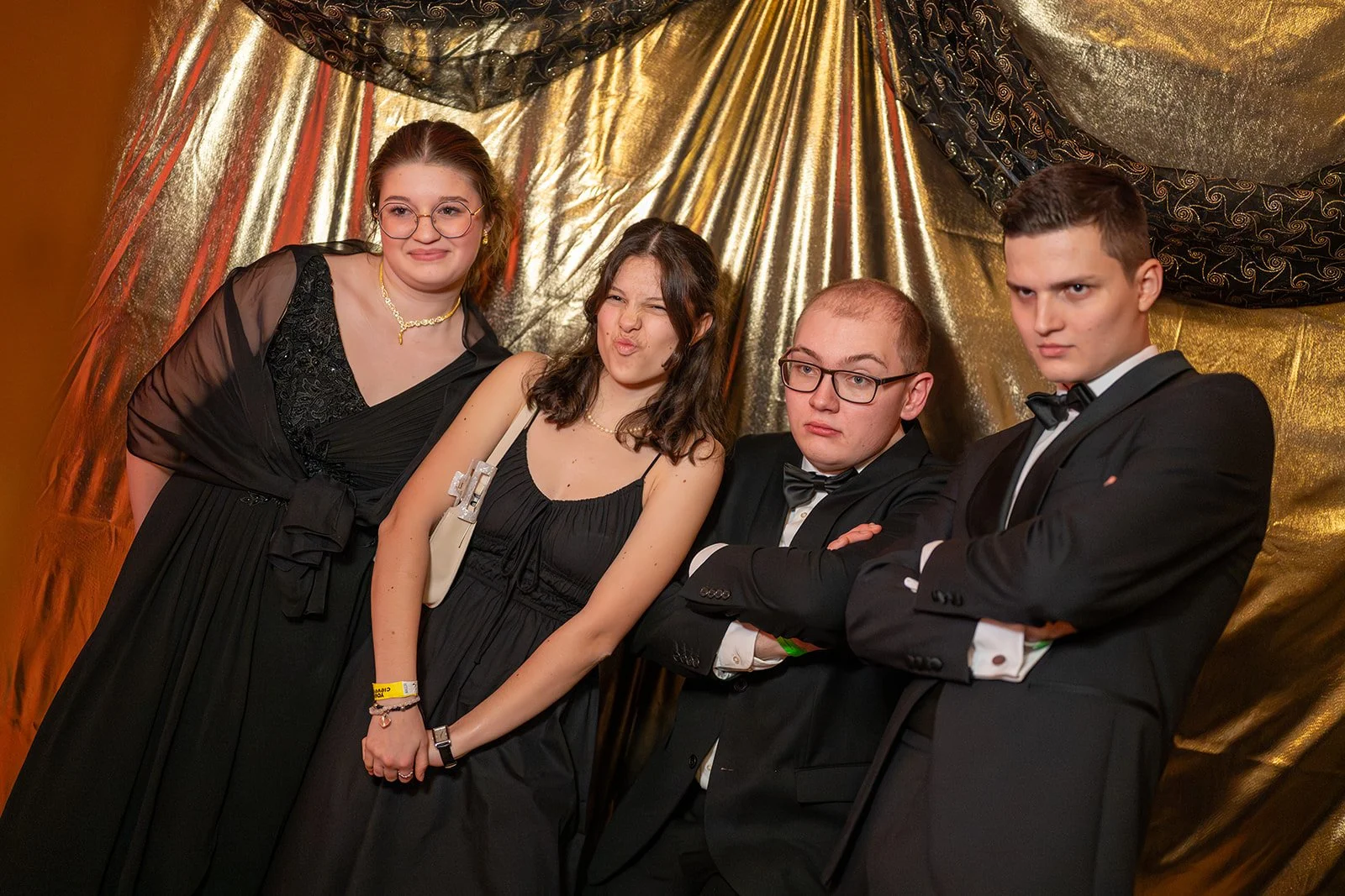 Vier junge Menschen in formeller Kleidung, zwei Frauen und zwei Männer, stehen vor einer goldfarbenen Dekoration. Die Frauen tragen schwarze Kleider, die Männer Anzüge mit Fliegen. Sie posieren in einer Gruppe, die dritte Person macht eine alberne Mi