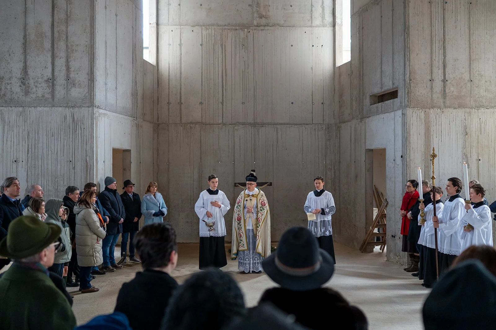 Ein religiöser Gottesdienst oder Zeremonie findet in einer rohen, unvollendeten Betonkirche statt. Ein Priester steht in der Mitte, umgeben von Ministranten und einer Gemeinde, die in der Kirche versammelt ist, einige in Herbst- und Winterkleidung.