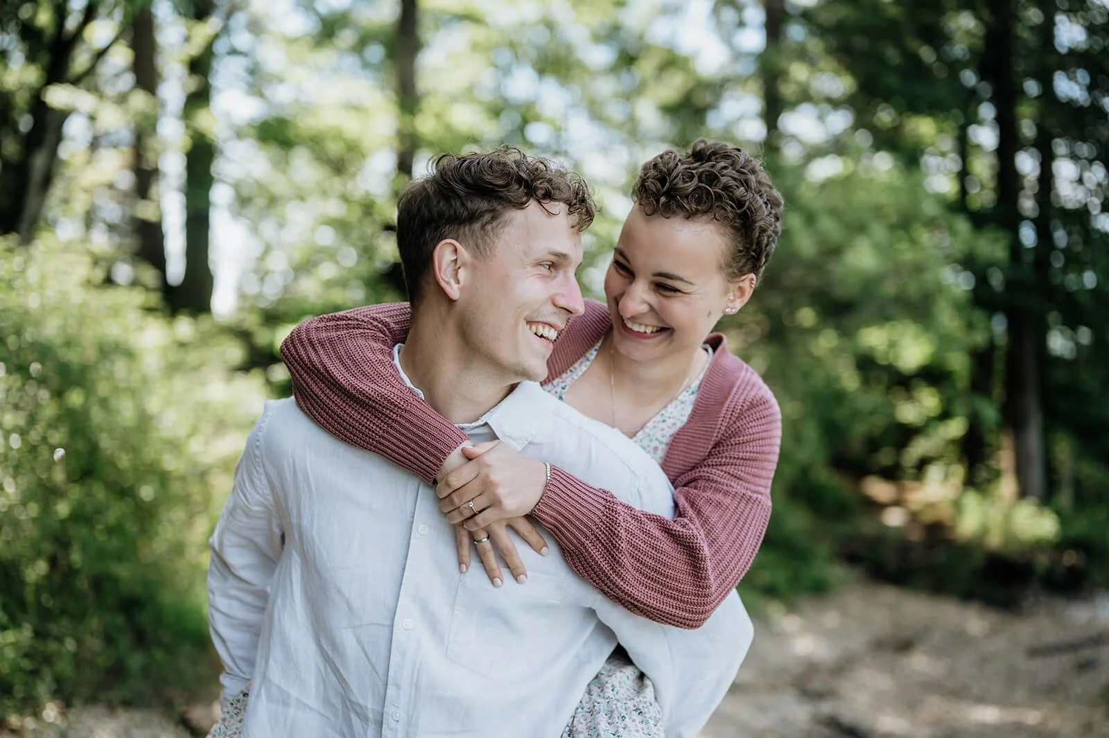 Ein lachendes Paar, der Mann trägt ein weißes Hemd, die Frau trägt eine rosa Strickjacke, beide im Grünen, bei Tageslicht.