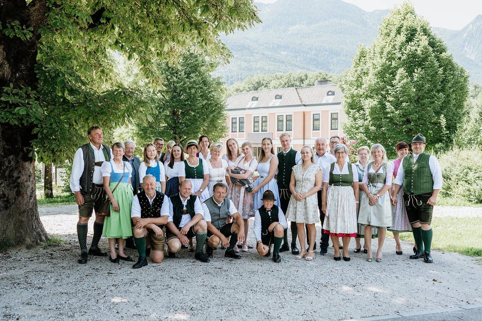 Gruppe von Menschen in traditionellen bayerischen Trachten im Freien, mit Bäumen und einem Gebäude im Hintergrund.