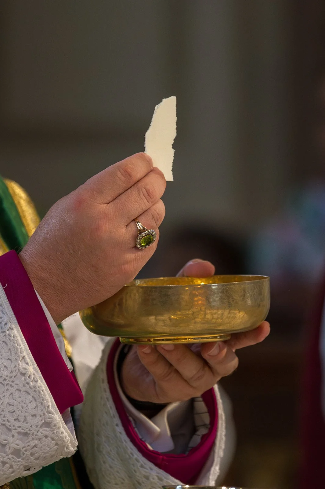 Eine Hand hält eine goldene Schale, die andere Hand hält ein zerfetztes Stück Papier. Die Person trägt einen großen Ring mit einem grünen Edelstein und ein Kleid mit weißen Lace-Ärmeln und pinker Einfassung.
