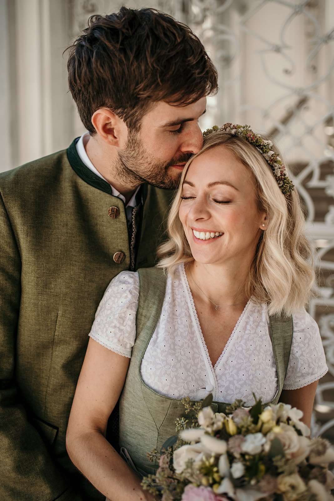 Ein glückliches Paar bei einer Hochzeit, der Mann küsst die Frau auf die Stirn, sie lächelt. Sie trägt ein Blumenkleid und eine Blumenkrone, er trägt eine traditionelle grüne Trachtenjacke.
