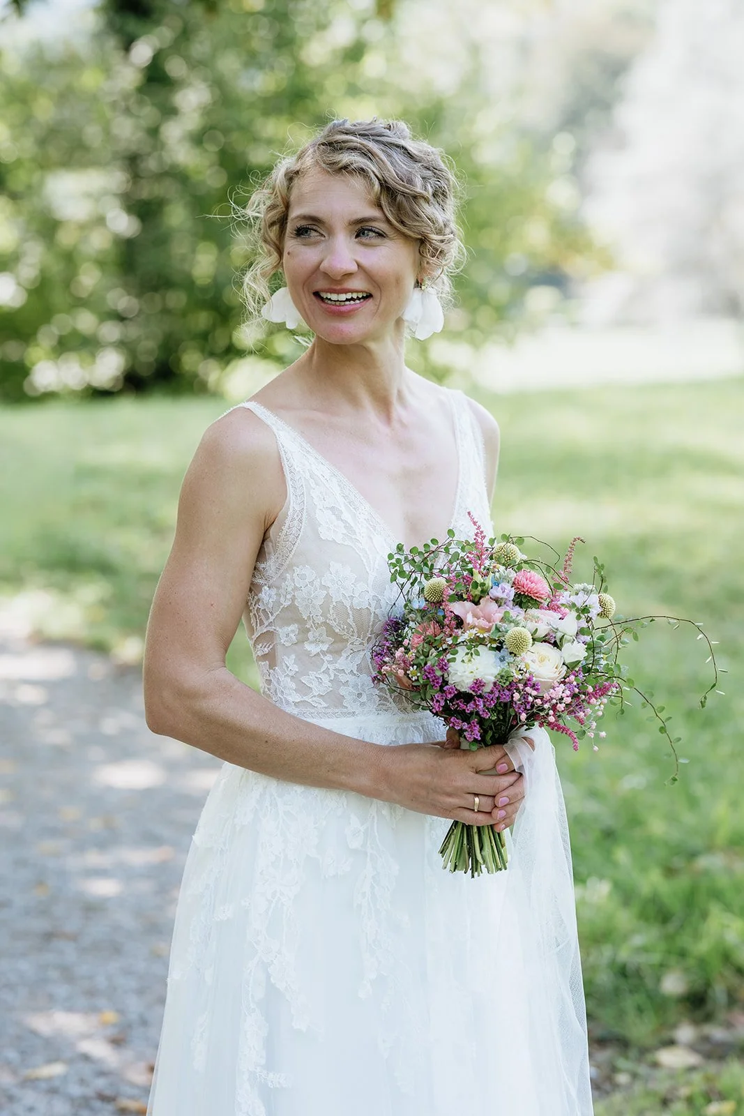 Eine Frau in einem weißen Brautkleid hält einen bunten Blumenstrauß im Freien, umgeben von grünen Bäumen.