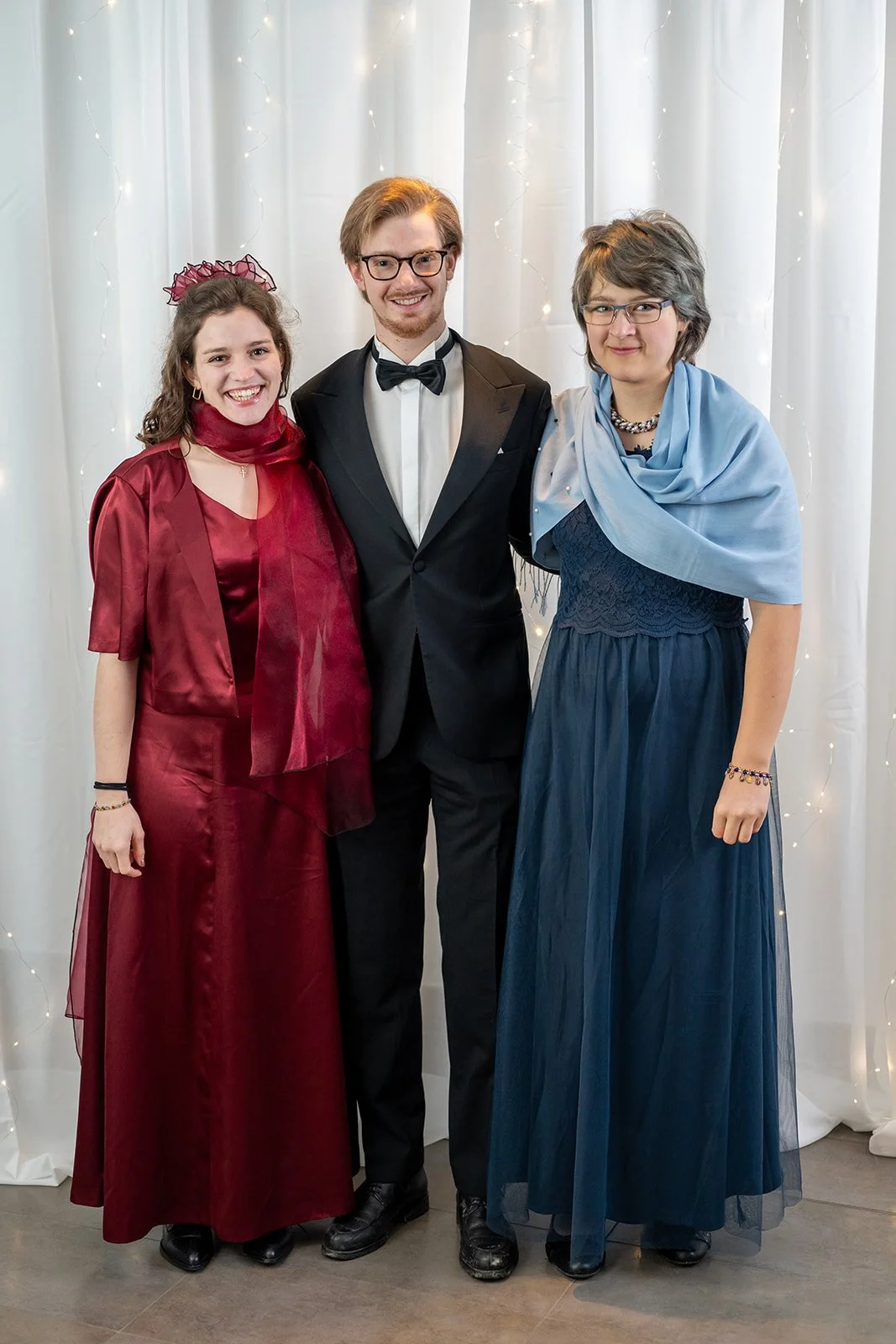Drei Menschen in formeller Kleidung stehen vor einem weißen Vorhang mit Lichterketten. Ein Mädchen in rotem Kleid und Haarreif, ein junger Mann in Anzug und Fliege, und ein Mädchen in blauem Kleid mit Schal.