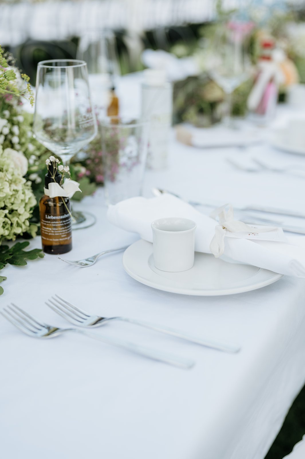 Ein elegant gedeckter Esstisch mit weißem Tuch, Wasser- und Weingläser, weißen Tellern, Besteck und einer weißen Tassen. Der Tisch ist mit Blumen und Dekorationen für eine Feier geschmückt.