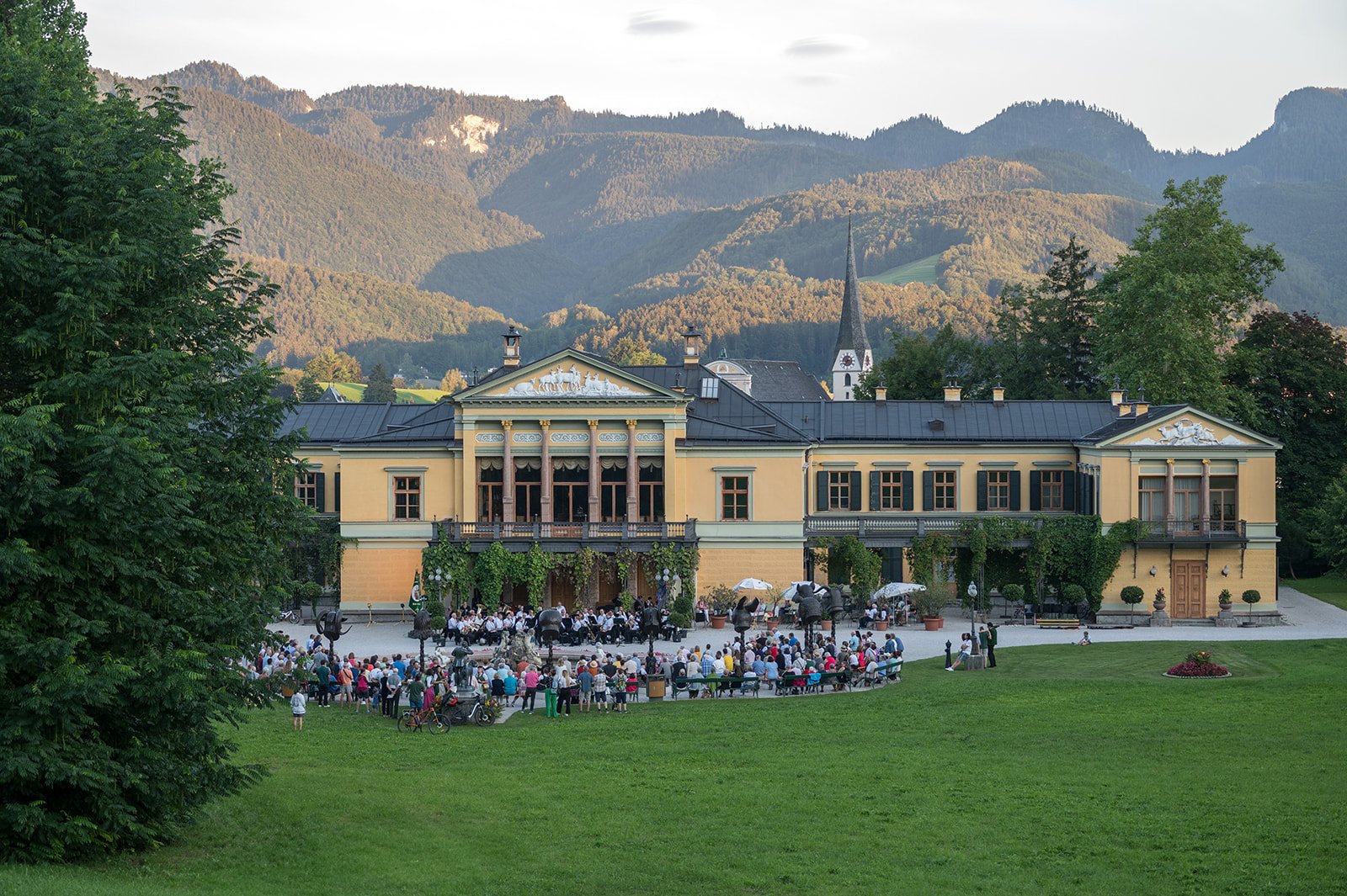 Historisches Gebäude mit einem großen Garten inmitten von Bergen, Menschen versammelt bei einem Event auf der Wiese.