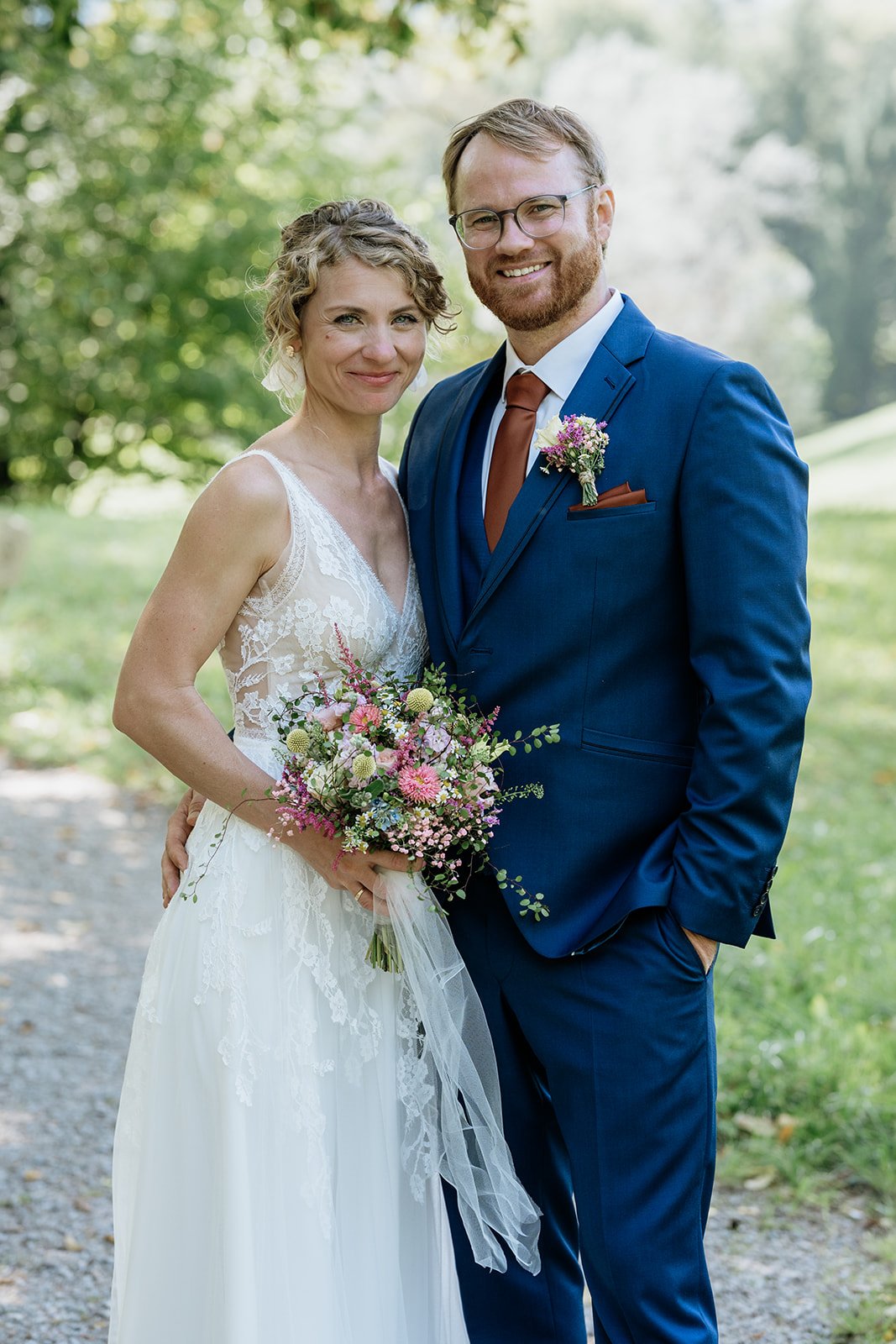 Ein Brautpaar steht draußen in einem Park, die Braut trägt ein weißes Hochzeitskleid, hält einen Blumenstrauß, und der Bräutigam trägt einen blauen Anzug mit braunem Krawatten-Accessoire.