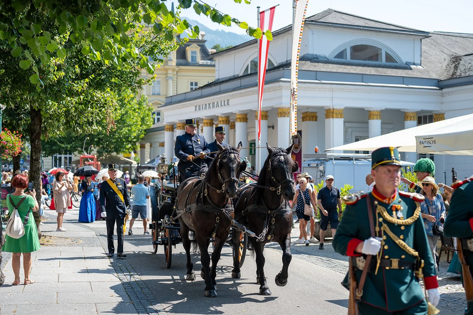 Historische Parade mit Kutschen und Soldaten in traditionellen Uniformen vor einem weißen Gebäude mit Säulen, umgeben von Menschen mit Sonnenschirmen an einem sonnigen Tag.