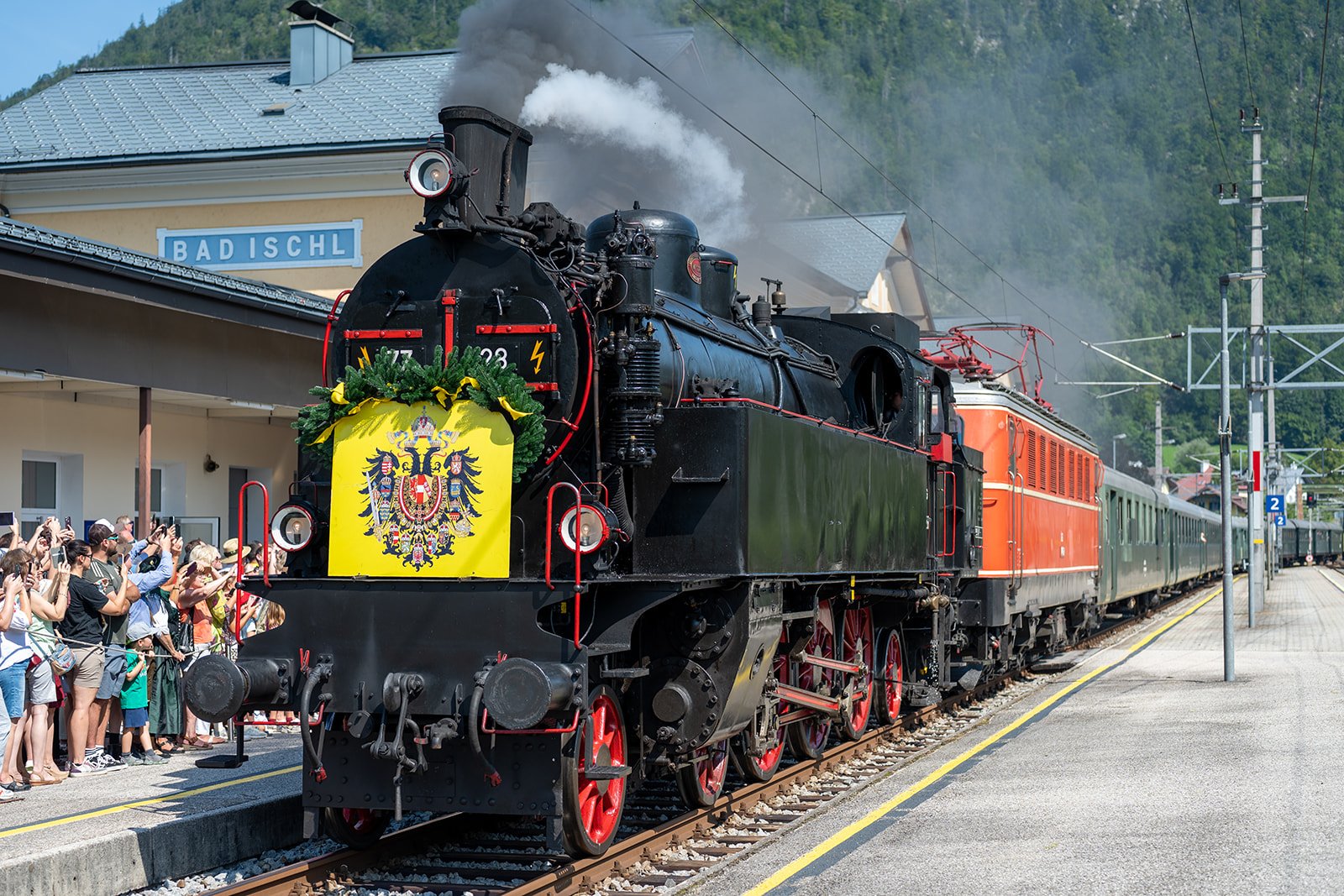 Eine schwarze Dampflokomotive mit roten Rädern und einer Deutschlandflagge vor einem Busbahnhof, während Menschen Fotos machen.