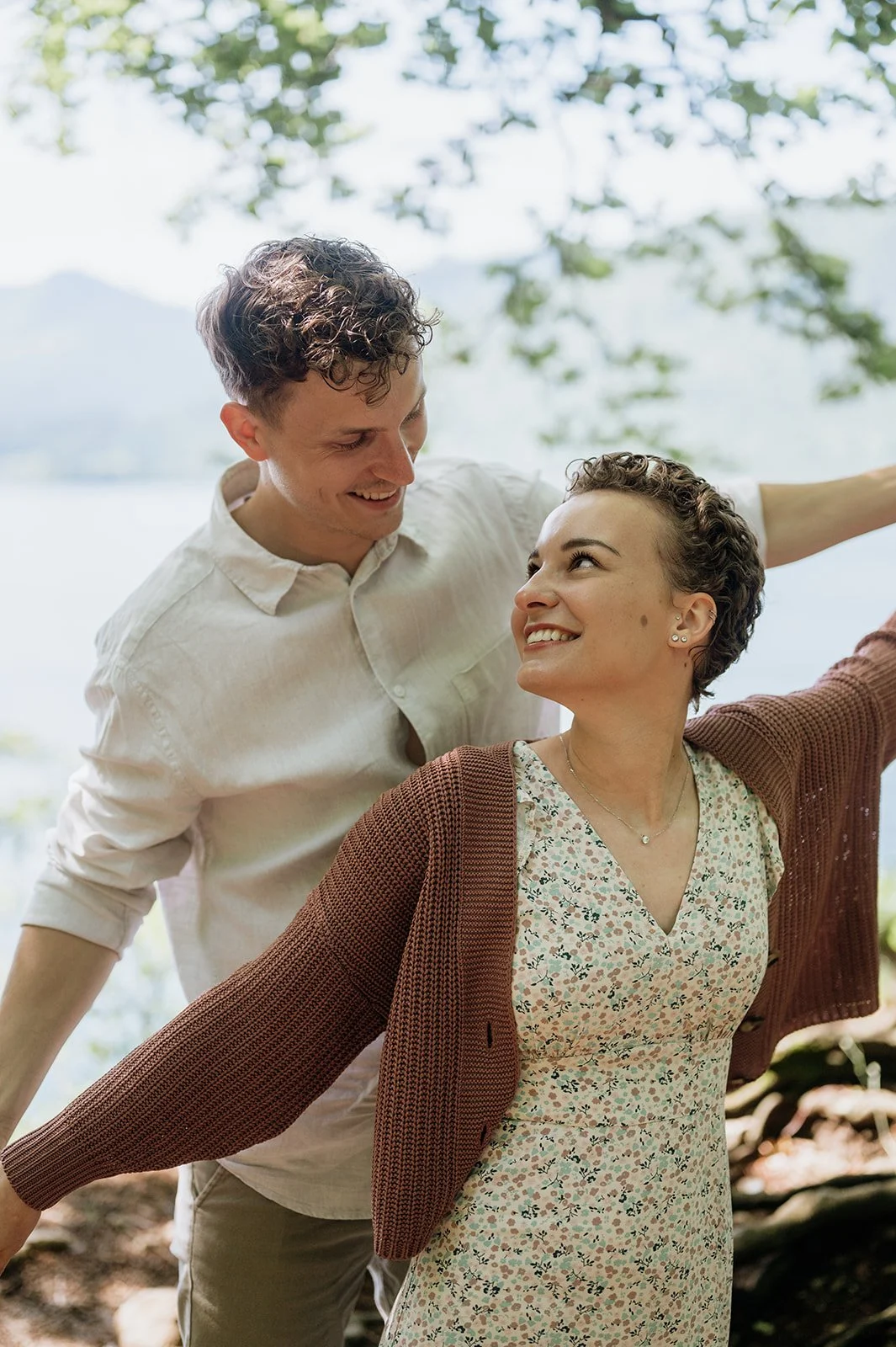 Ein lachendes Paar, das im Freien am Wasser steht, umgeben von Bäumen, bei Sonnenschein.
