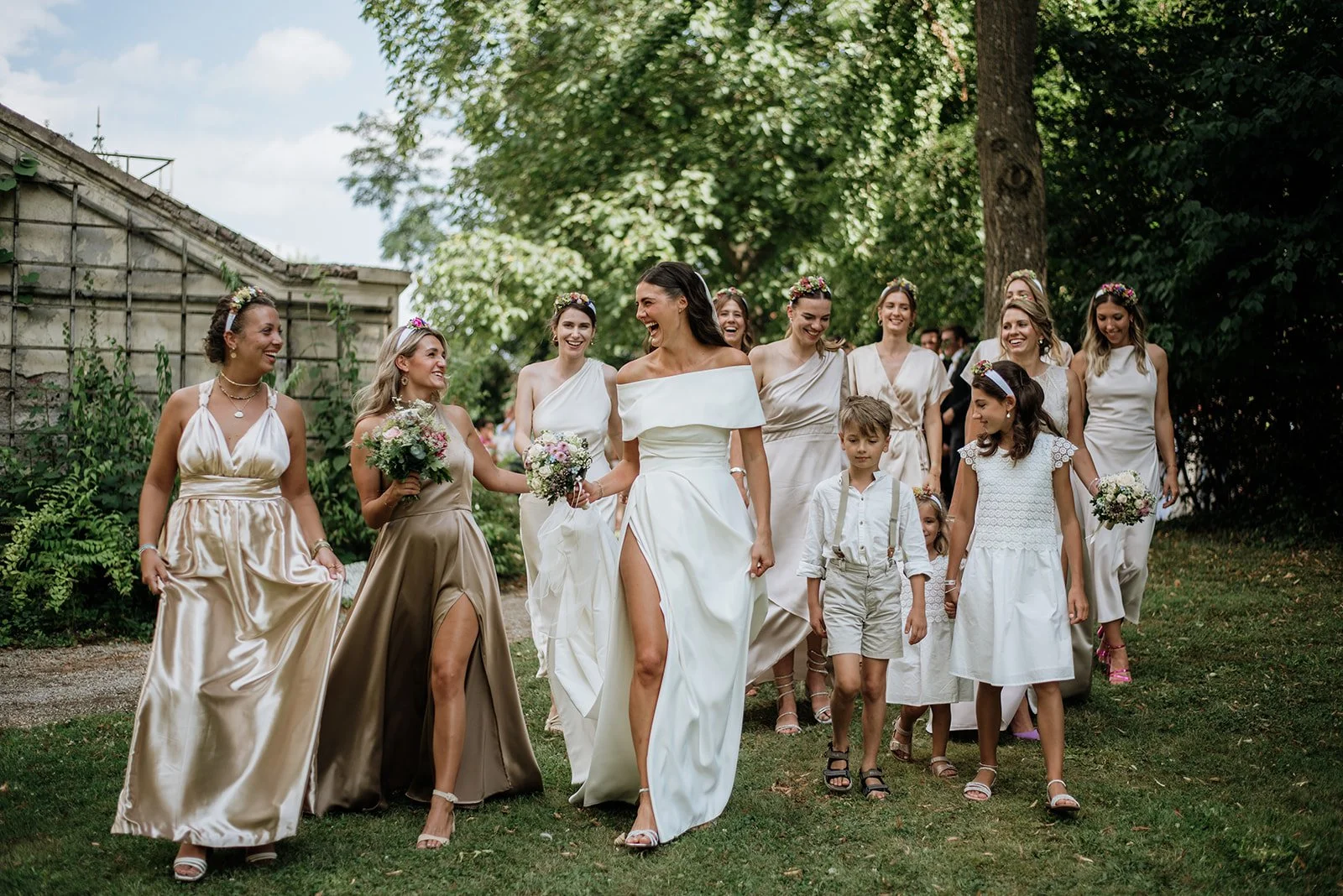 Hochzeitspaar mit Brautjungfern und Kindern beim Spaziergang im Garten, lachend und in eleganten Kleidern, mit Blumen in der Hand.