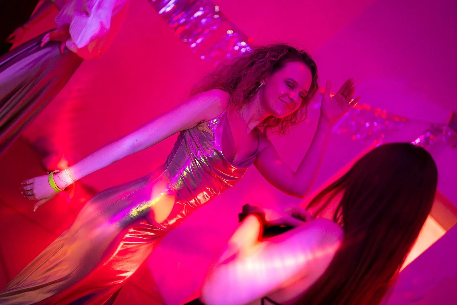 Zwei Frauen tanzen in einem Raum mit pinker und lila Beleuchtung, eine trägt ein metallisches, glänzendes Kleid, die andere hat lange dunkle Haare.