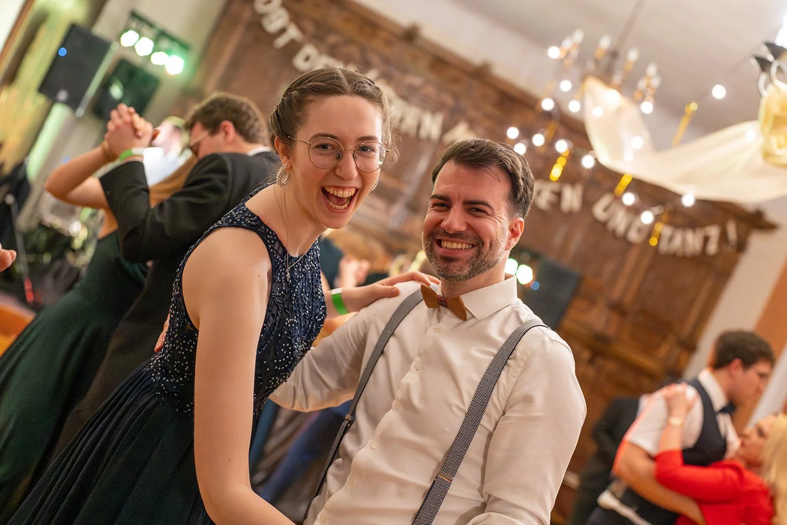 Zwei lachende Menschen, ein Mann mit Hemd, Hosenträger und Fliege, eine Frau mit Brille und eleganten Kleid, tanzen auf einer Party in einem festlich dekorierten Raum.