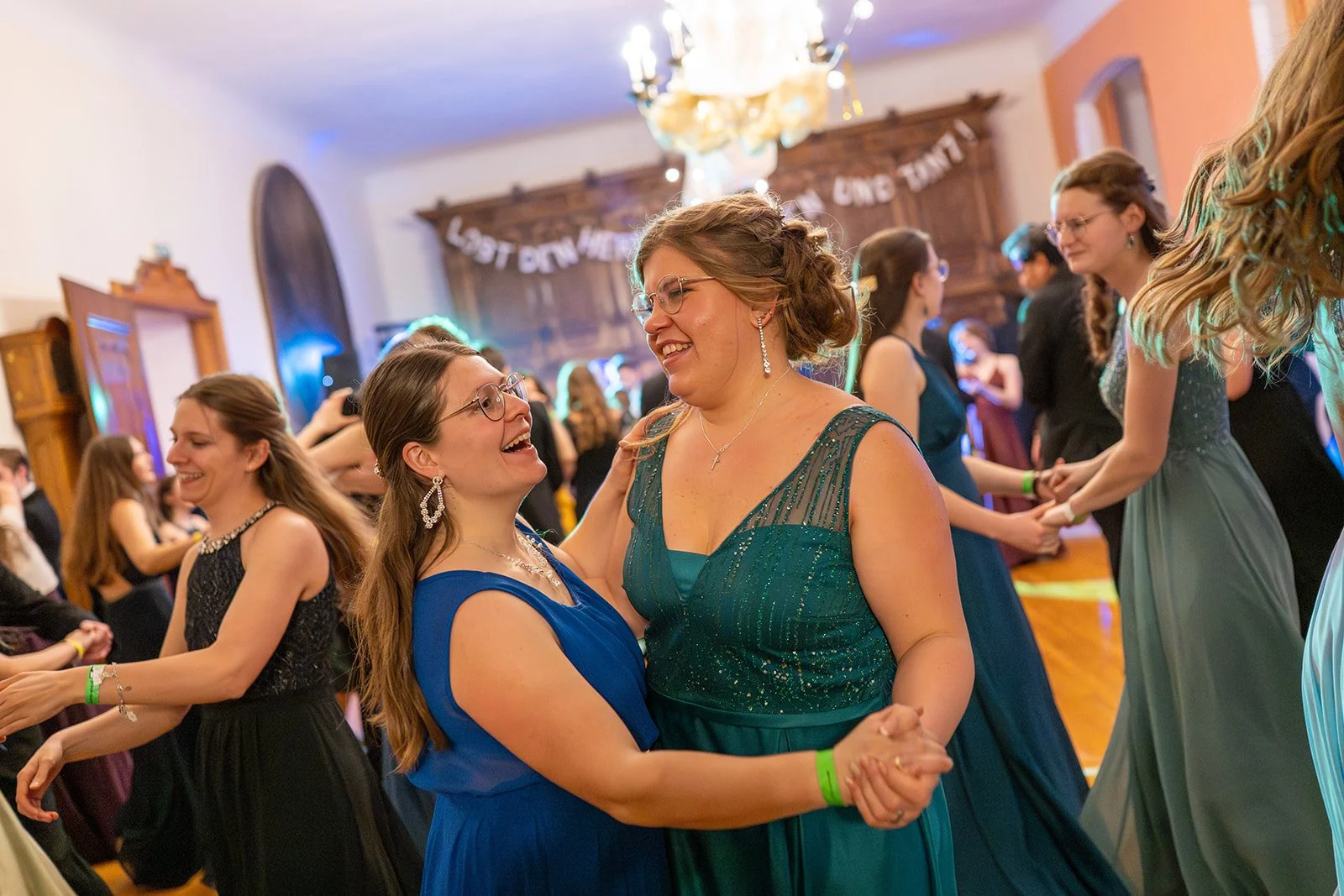 Frauen tanzen auf einer Feier im Festsaal, lächeln, in eleganten Abendkleidern, festliche Stimmung, buntes Dekor im Hintergrund.