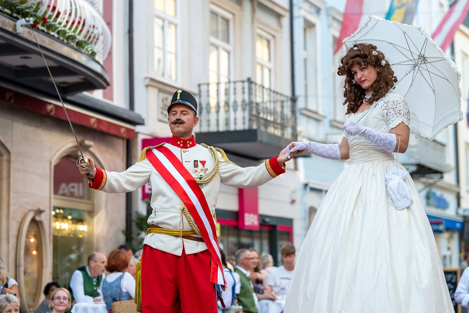 Ein Mann in historischen Militäruniformen tanzt mit einer Frau in einem weißen Ballkleid bei einem Straßenfest.