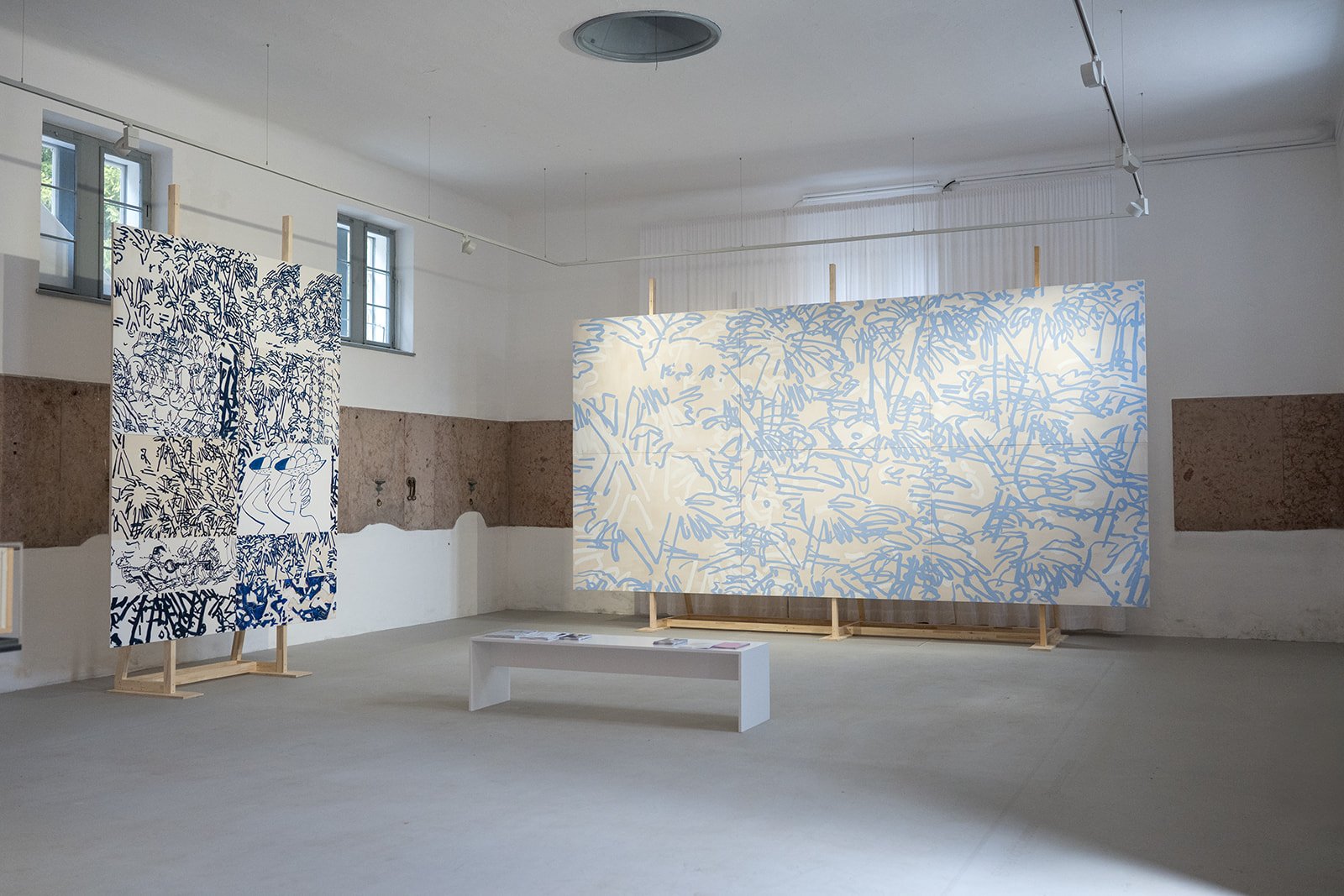 Kunstinstallation mit zwei großen Wandpanelen mit blauen Linienzeichnungen auf weißem Hintergrund in einem Raum mit weißen Wänden, kleinen Fenstern und Betonboden.