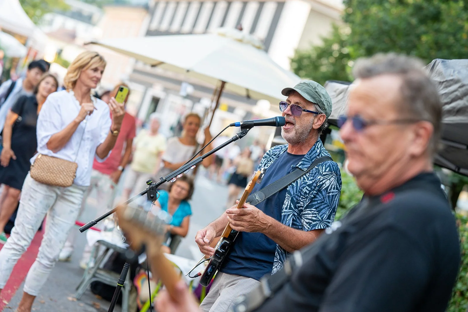Ein Mann mit Gitarre singt und spielt Musik auf einer Straße, umgeben von Menschen, die zusehen und Fotos machen.