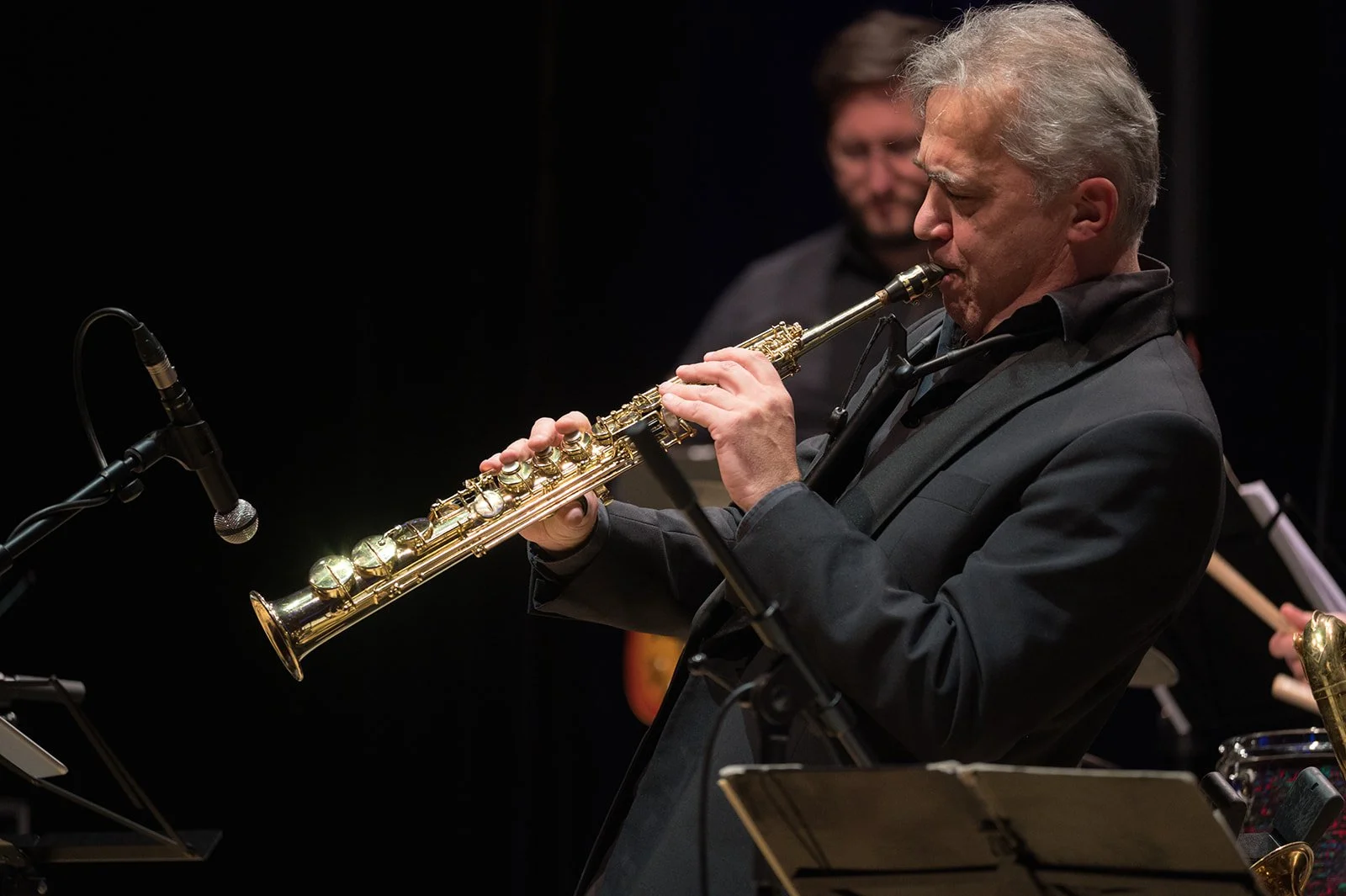 Ein Mann spielt auf einem Sopranino-Saxophon bei einem Auftritt, im Hintergrund ist eine andere Person mit einem Musikinstrument zu sehen.