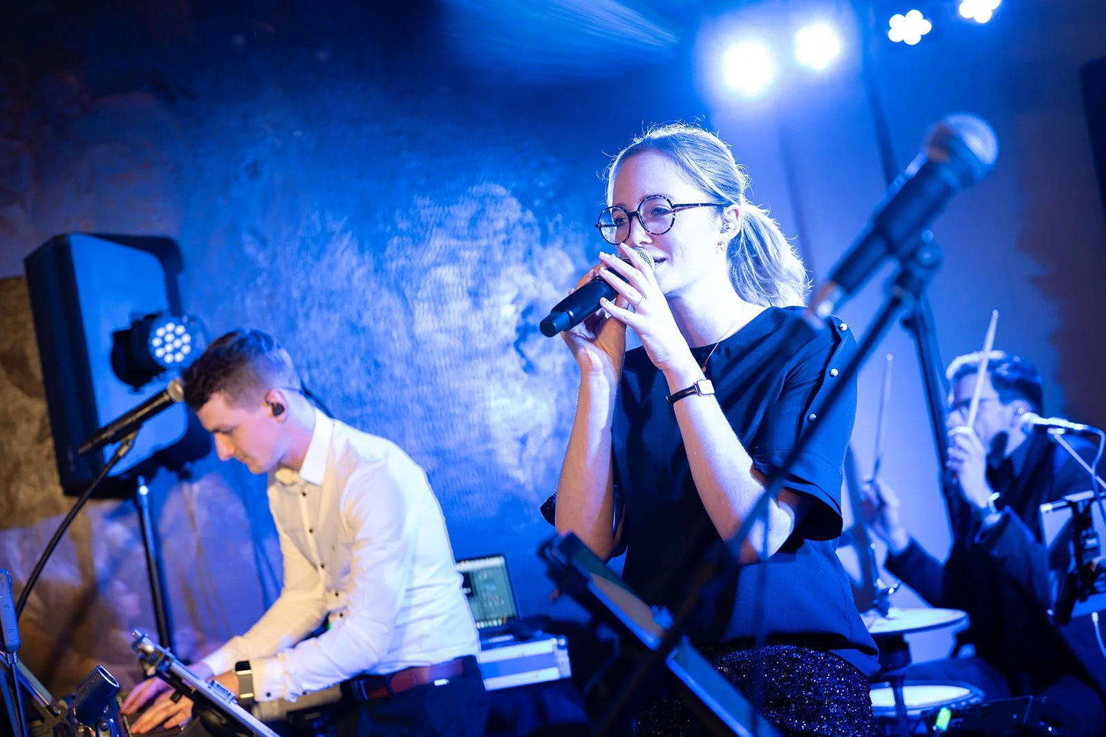 Eine junge Frau mit Brille singt mit einem Mikrofon auf einer Bühne, während zwei Musiker im Hintergrund ein Klavier und ein Instrument spielen, bei einer Live-Performance unter blauen Scheinwerfern.