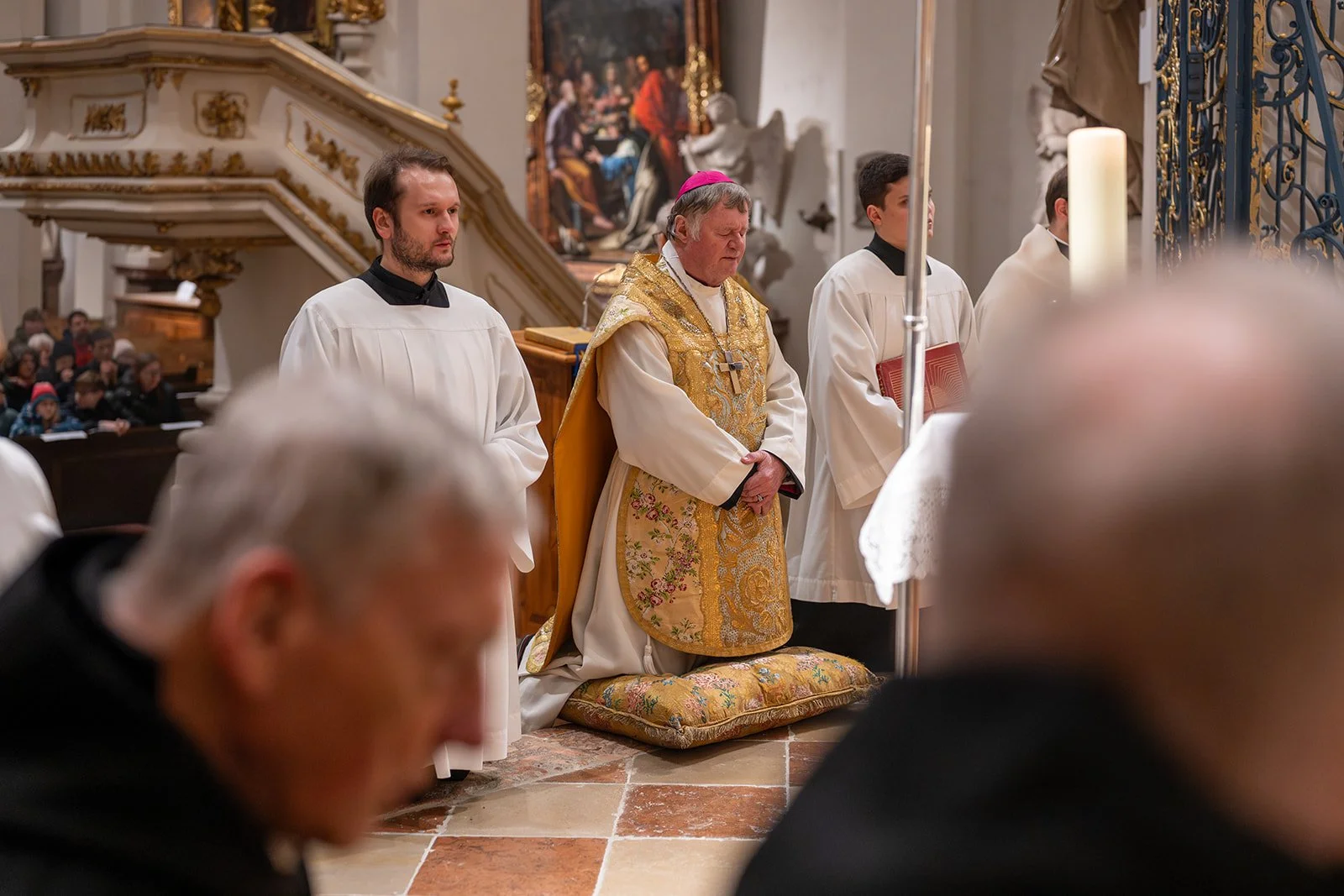 Ein katholischer Priester und drei Ministranten während eines Gottesdienstes in einer Kirche, mit angeschlossener Menschenmenge im Hintergrund.
