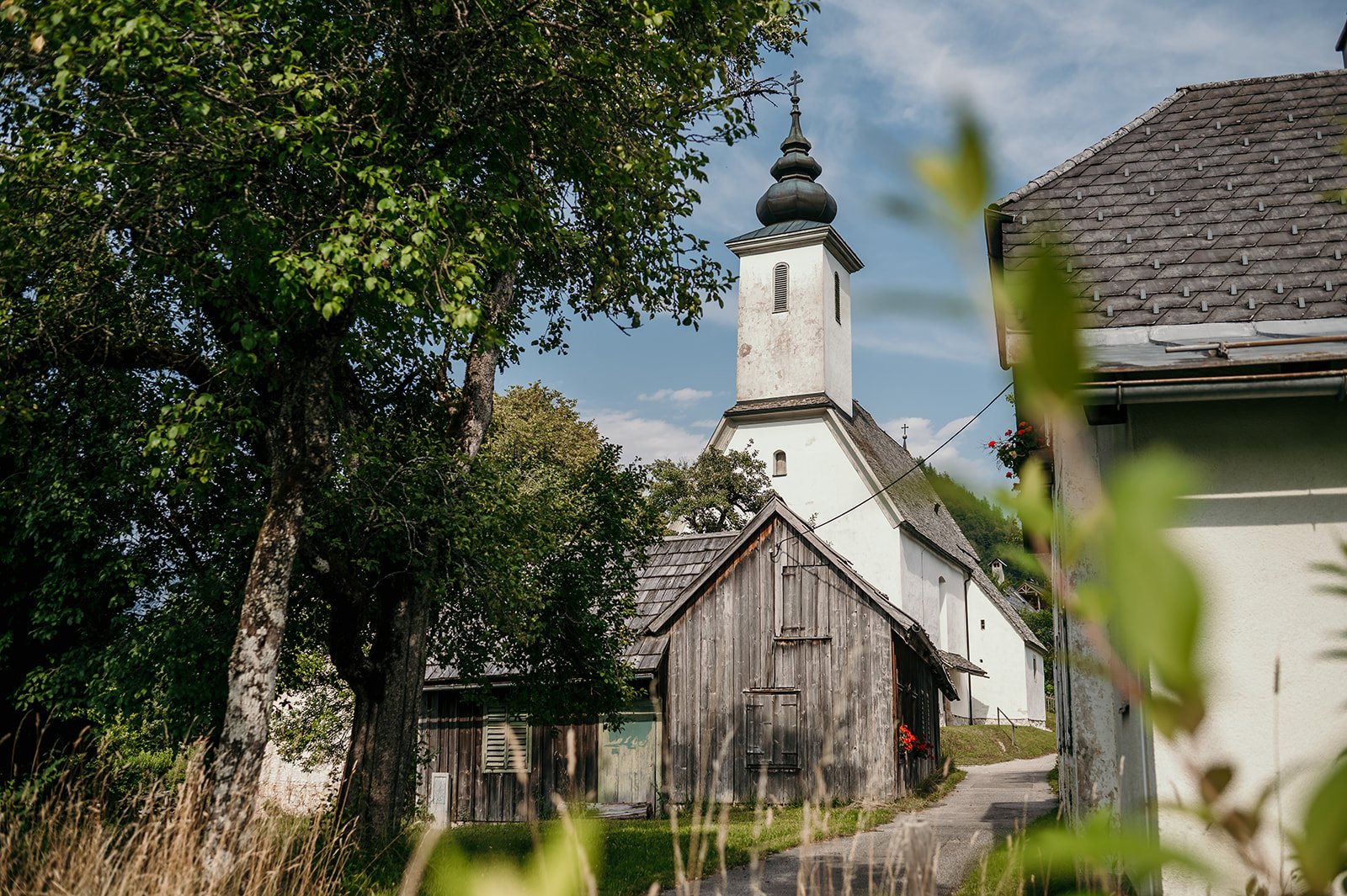 Eine kleine Kirche mit einem schwarzen Dach und weißen Wänden, umgeben von Bäumen und Holzhäusern auf einem ländlichen Weg.
