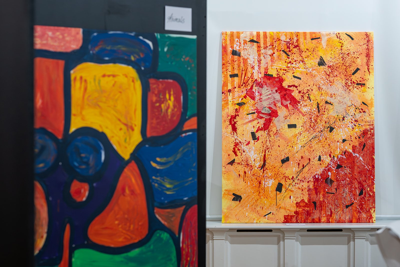 Zwei abstrakte Gemälde, eines im Vordergrund mit großen, bunten geometrischen Formen und das andere im Hintergrund mit roten, orangefarbenen und schwarzen Flecken und Linien, in einer Kunstgalerie