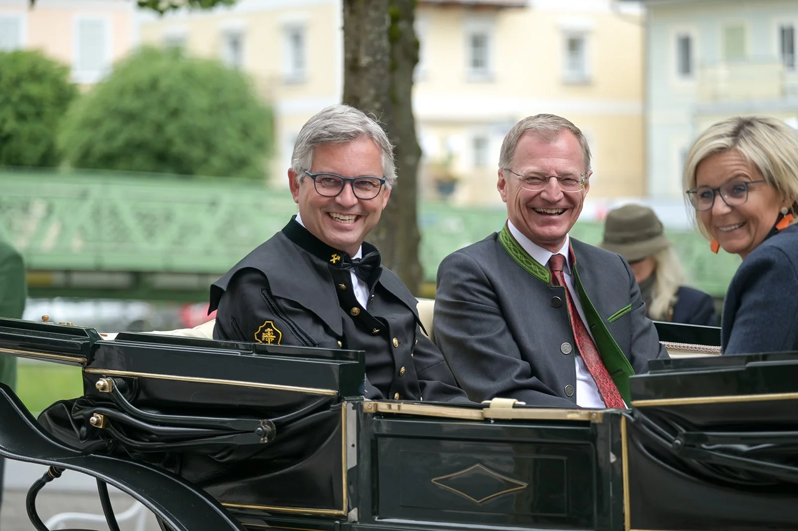 Drei Menschen in traditionellen Trachten sitzen in einer Kutsche und lachen. Im Hintergrund sind Bäume und mehrstöckige Gebäude sichtbar.
