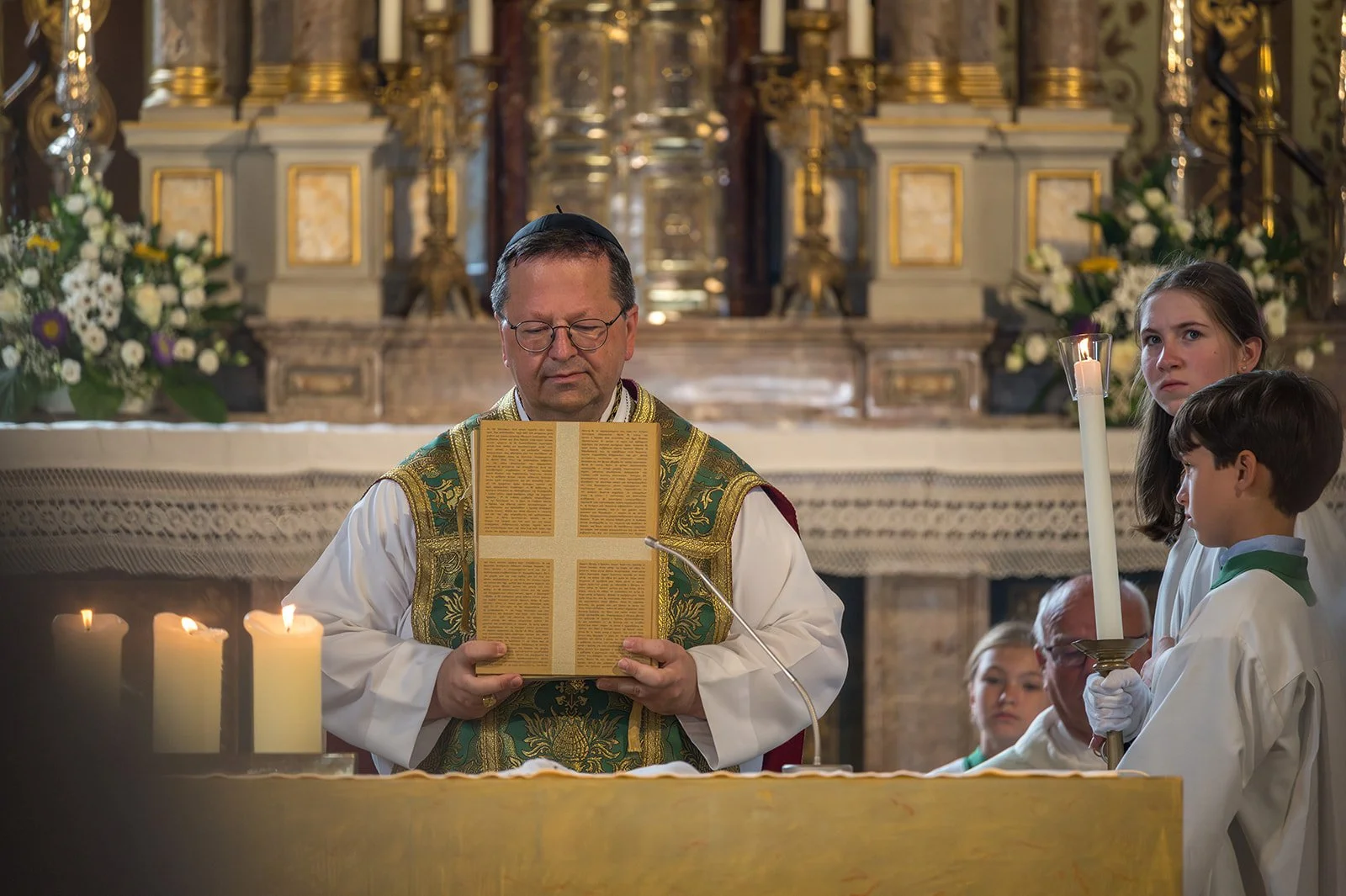 Priester steht in einer Kirche mit geöffnetem Buch, umgeben von Kerzen und Blumentöpfen, während er eine Zeremonie durchführt. Zwei Kinder, ein Mädchen und ein Junge, halten eine große Kerze, sie tragen weiße Gewänder, im Hintergrund sind weitere Per