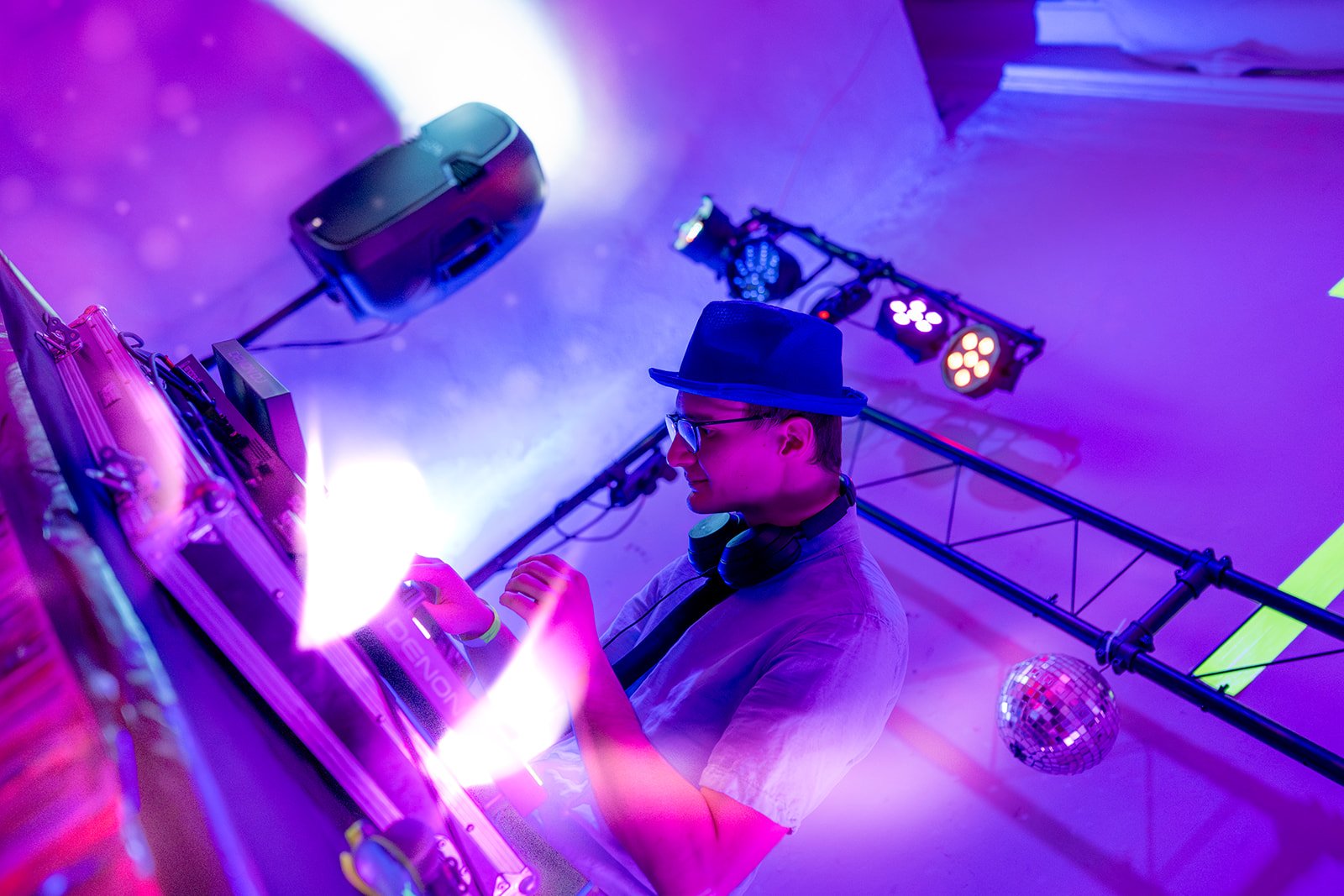 Ein junger DJ mit Hut, Brille und Kopfhörern, der an einem Mischpult bei einer Veranstaltung auflegt, umgeben von buntem Licht und Discokugeln.