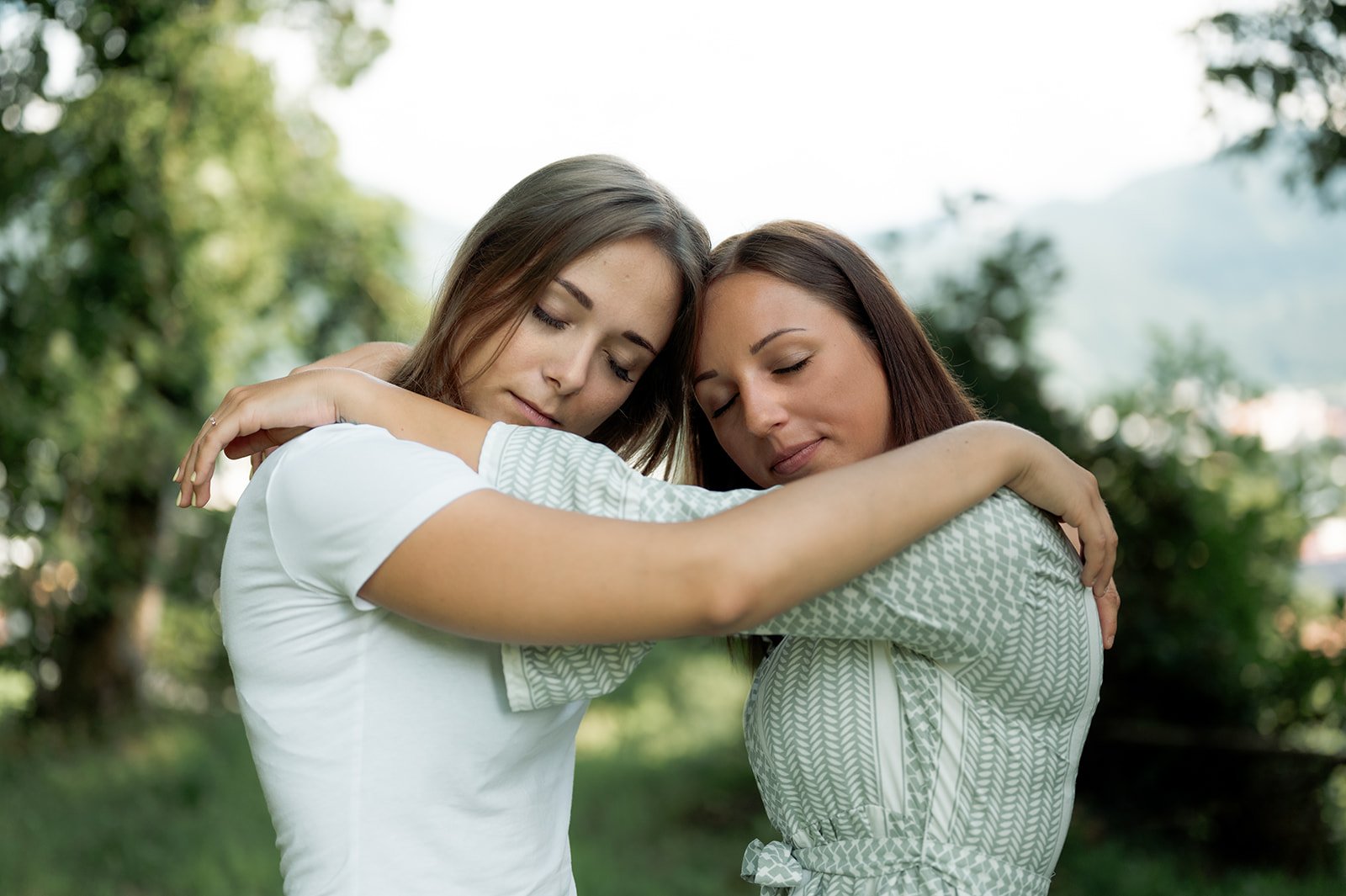 Zwei Frauen umarmen sich in einem grünen Park, mit entspannten Gesichtern und geschlossenen Augen.