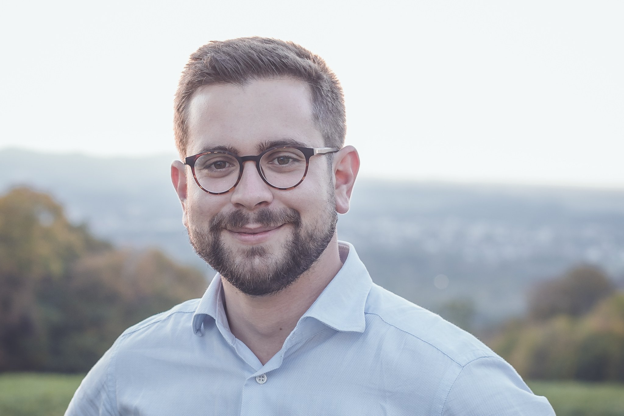 Porträt eines lachenden Mannes mit Brille, Bart und kurzen Haaren, stehend im Freien mit einem ländlichen Hintergrund und Blick auf die Landschaft.