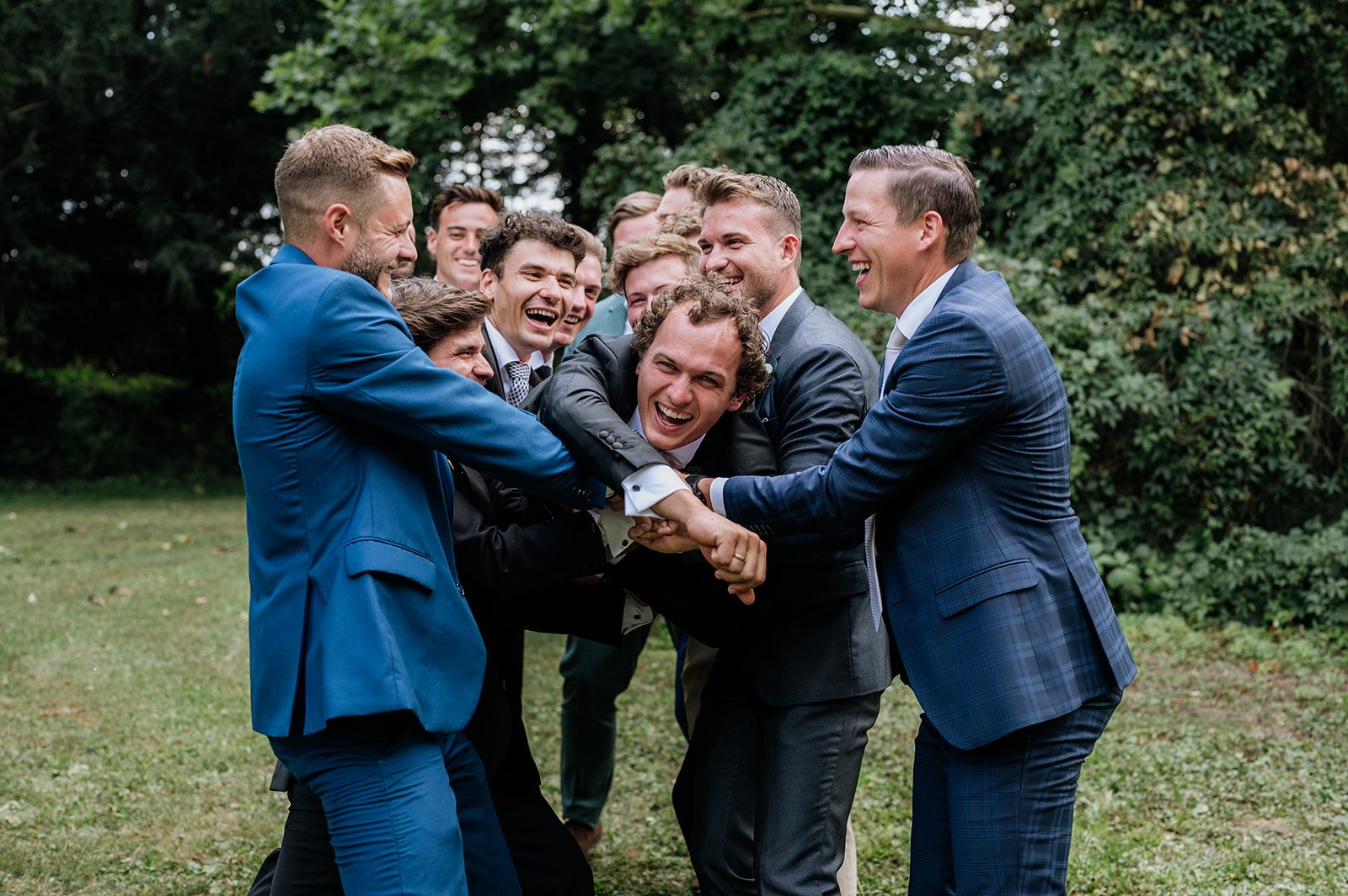 Männer in formeller Kleidung, die sich spielerisch und lachend auf einer Wiese im Freien umarmen und an den Händen halten.