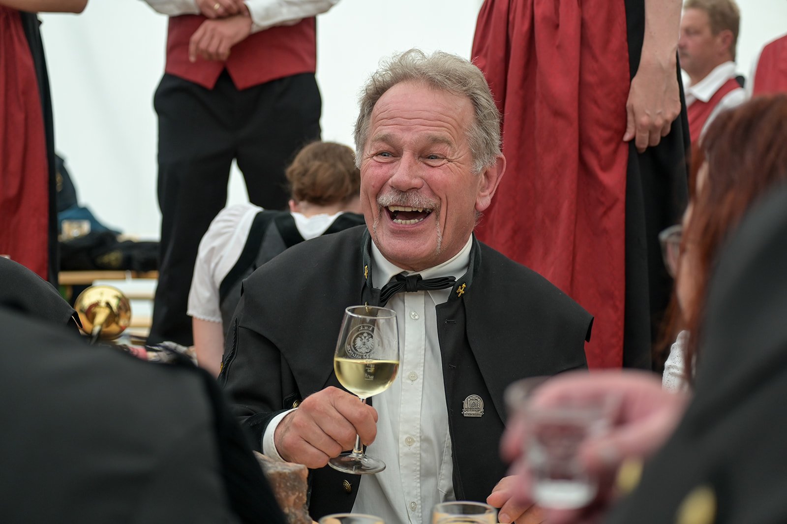 Ein lachender Mann in formeller Kleidung, der ein Glas Weißwein hält, bei einer Feier im Festzelt, hinter ihm sind weitere Gäste und Musikanten in traditionellen Uniformen.