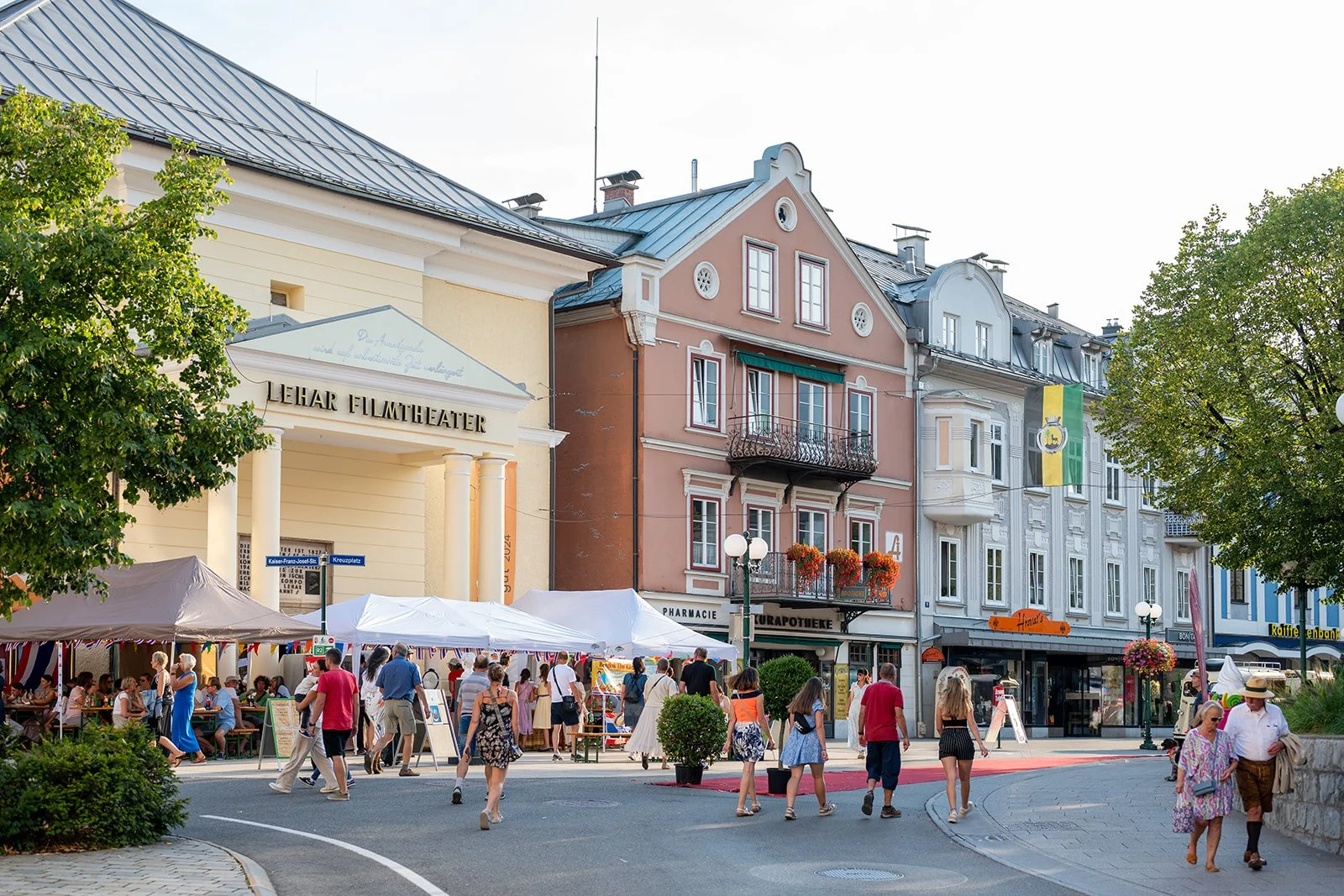Blick auf eine belebte Straße mit Menschen, Verkaufsständen und historischen Gebäuden in einer europäischen Stadt, mit einem Theater namens 'Lehar Filmtheater' im Vordergrund.