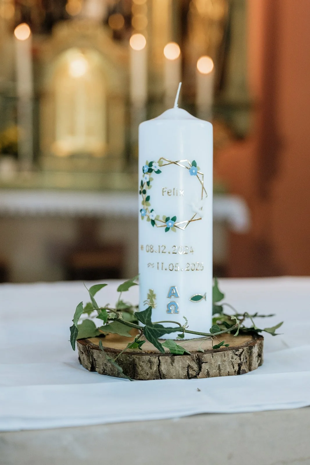 Weißer Kerze mit blauen Blumen, goldenen Akzenten und Texten, auf einem Holzscheit mit Blättern, im Hintergrund eine Kirche mit Kerzen.