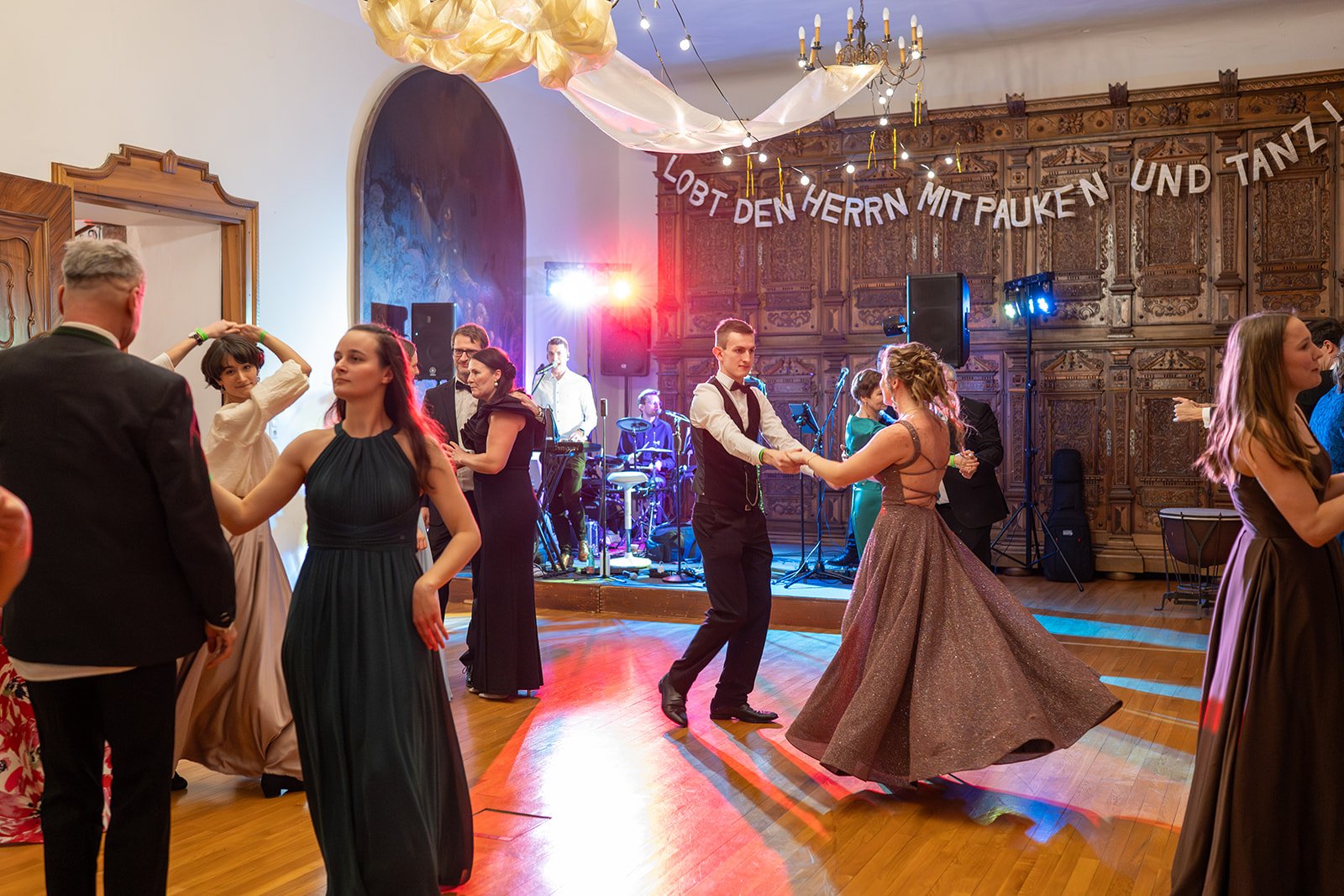 Menschen tanzen in eleganter Kleidung bei einer Feier in einem großen Saal, während eine Live-Band auf einer Bühne im Hintergrund spielt. An der Wand hängt ein Banner mit dem Text 'Lobet den Herrn mit Pauken und Tanzen'.