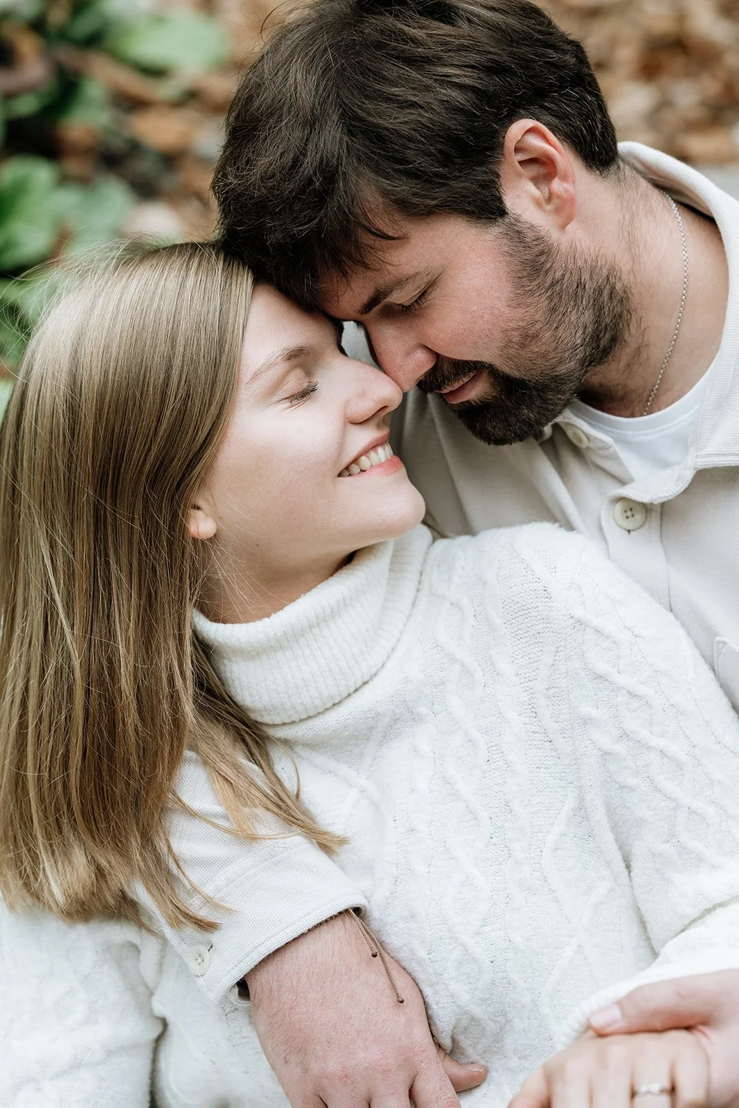 Ein Paar, das sich liebevoll gegenübersteht, sitzt im Freien auf dem Boden, umgeben von Laub und grünen Pflanzen.