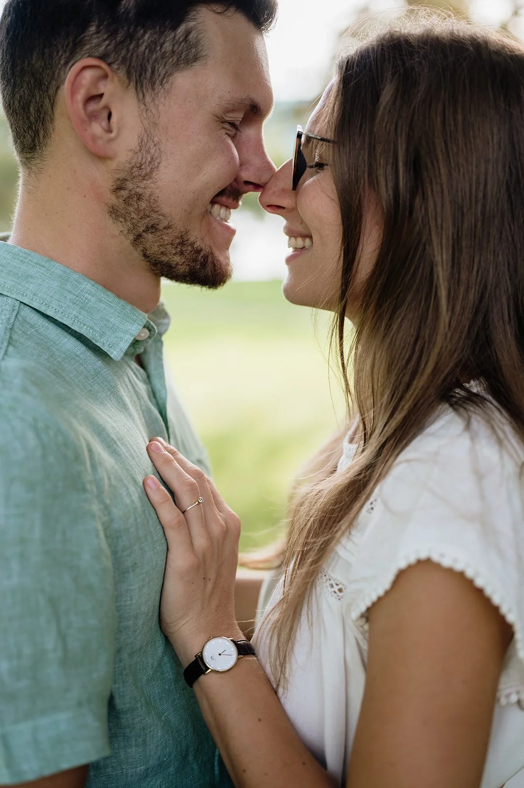 Ein Paar steht sich mit ihren Nasen berührend, lächelnd und sehr nah beieinander im Freien, die Frau trägt eine Armbanduhr und einen Ring.