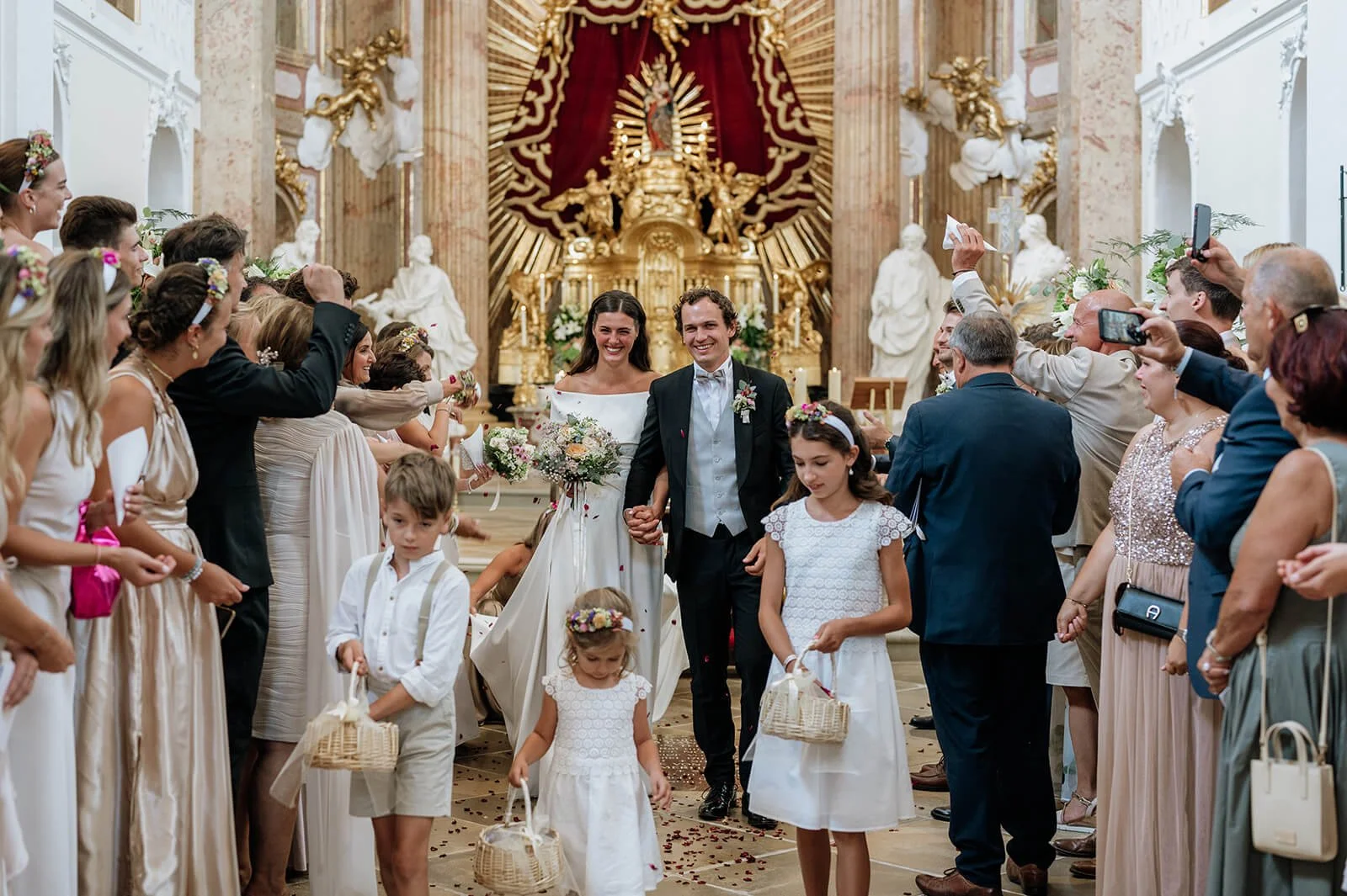 Ein Brautpaar läuft in einer Kirche Hand in Hand, umgeben von Gästen, die Fotos machen. Die Kirche ist reich mit goldenen Verzierungen und Skulpturen geschmückt.
