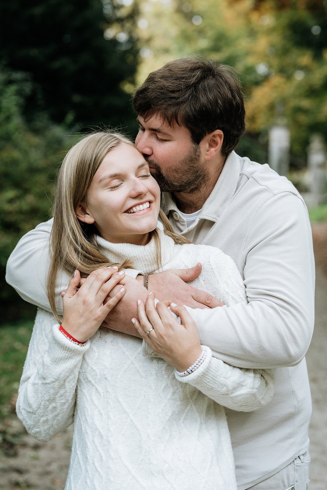 Ein lachendes junges Paar umarmt sich im Park, der Mann küsst die Frau auf die Stirn.