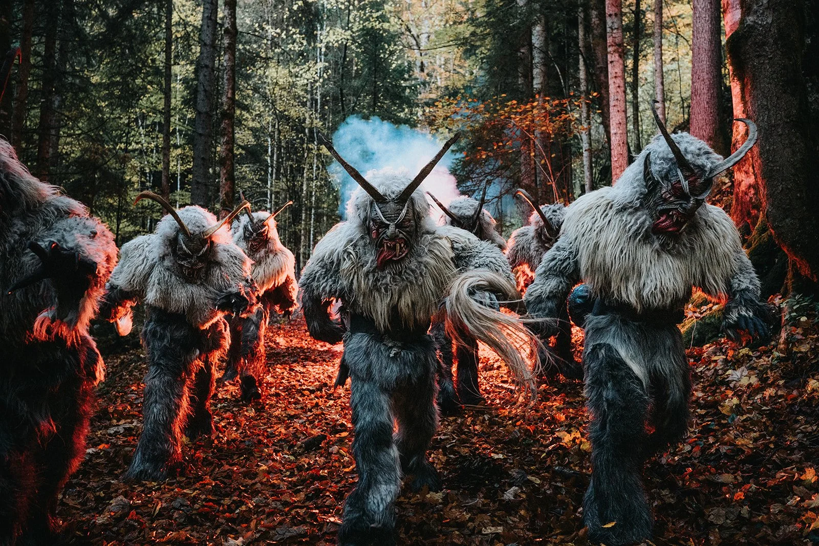 Eine Gruppe von fabelhaften, haarigen Kreaturen mit Hörnern und zerrissenen Gesichtern läuft durch einen dunklen, bewaldeten Pfad bei Sonnenuntergang oder Sonnenaufgang. Die Kreaturen haben ein unheimliches, mystisches Aussehen, beleuchtet von rotem 