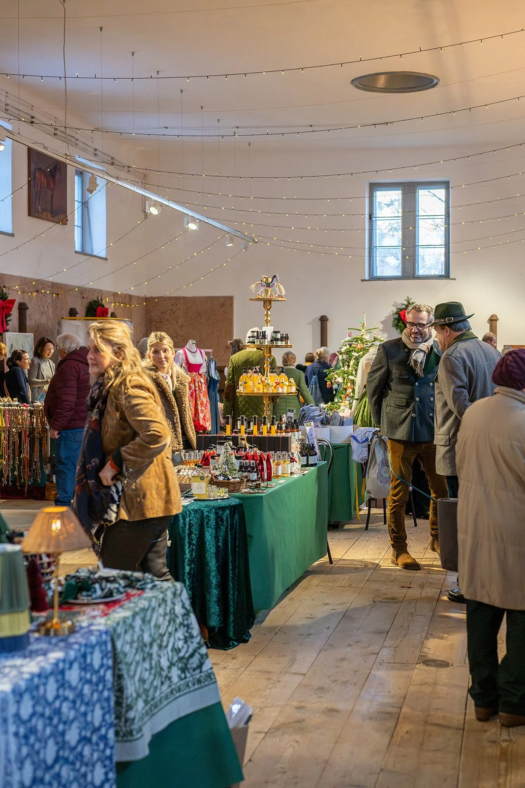 Blick in einen festlich geschmückten Marktstand in einem großen Saal mit Kunden, die Einkäufe betrachten. Der Saal hat hohe Fenster und ist mit Lichterketten dekoriert.