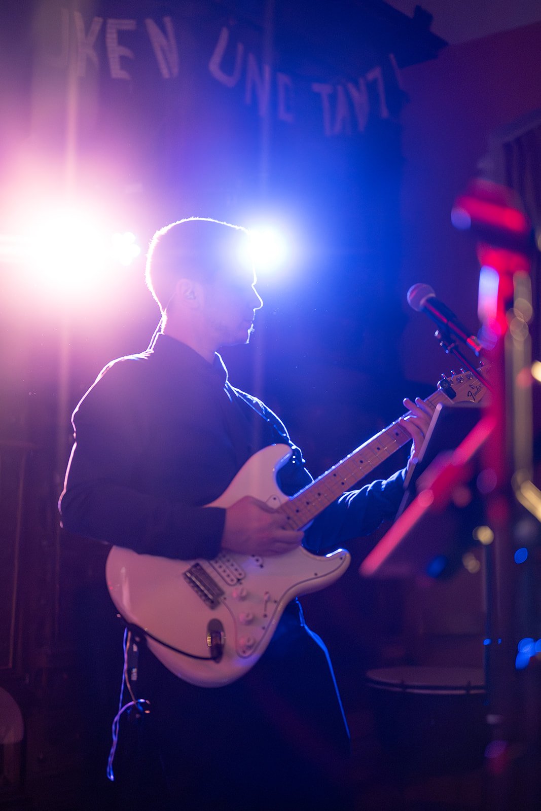 Ein junger Mann spielt Gitarre auf einer Bühne bei einem Live-Auftritt, im Hintergrund bunte Lichter und eine Dekoration mit Buchstaben