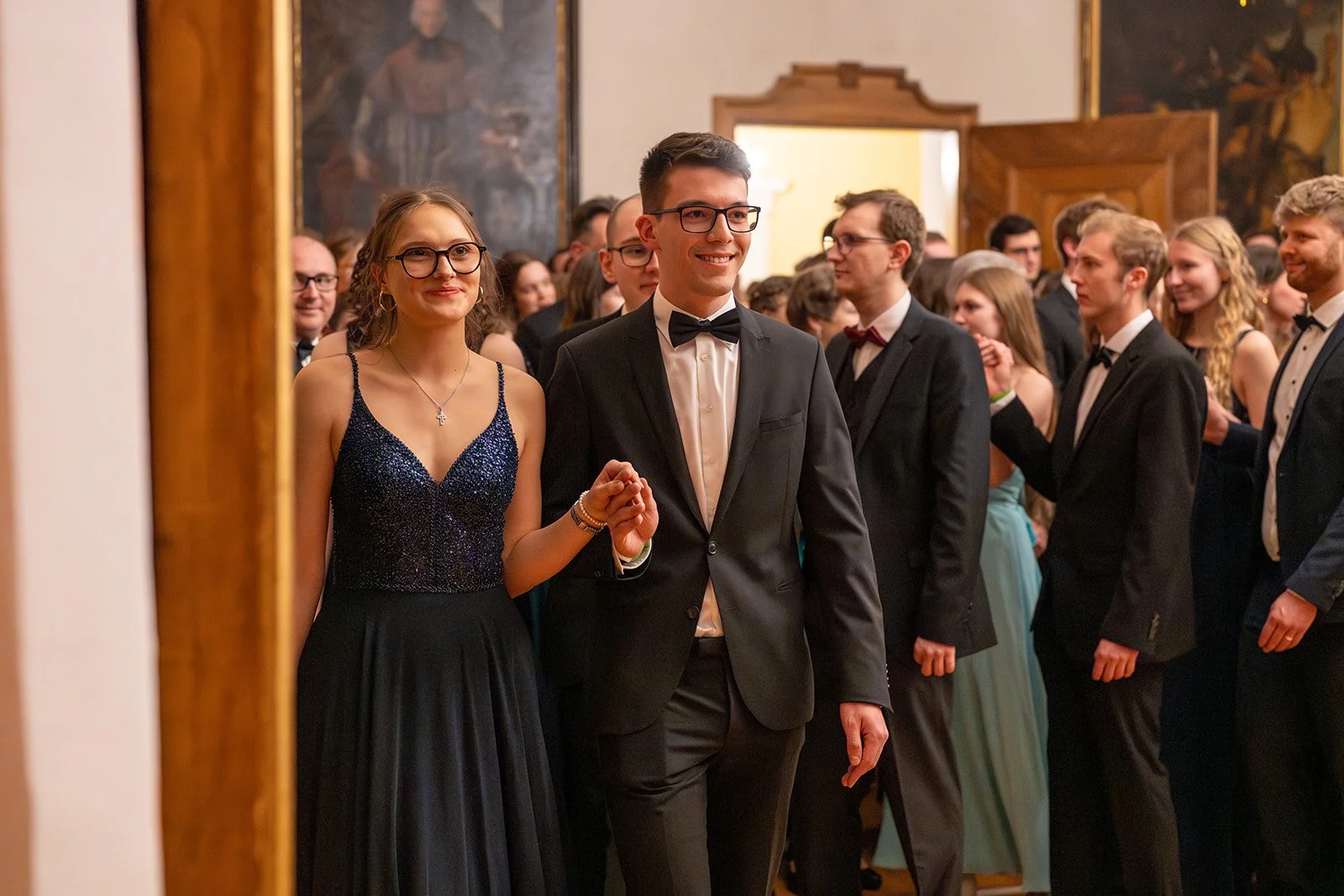 Junge Menschen in formeller Kleidung, die an einer Gala oder einem Ball teilnehmen, halten Händchen beim Tanzen, im Hintergrund befinden sich weitere Gäste und festliche Dekorationen.