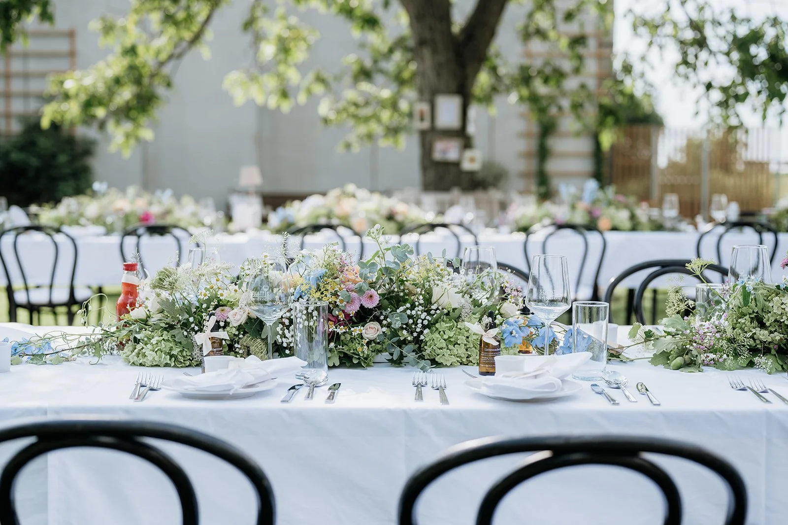 Ein festlich gedeckter Tisch im Freien mit weißen Tischtüchern, zahlreichen Blumenarrangements, leeren Weingläsern, Wasserflaschen und Besteck, umgeben von schwarzen Stühlen, bei sonnigem Wetter in einem Garten.